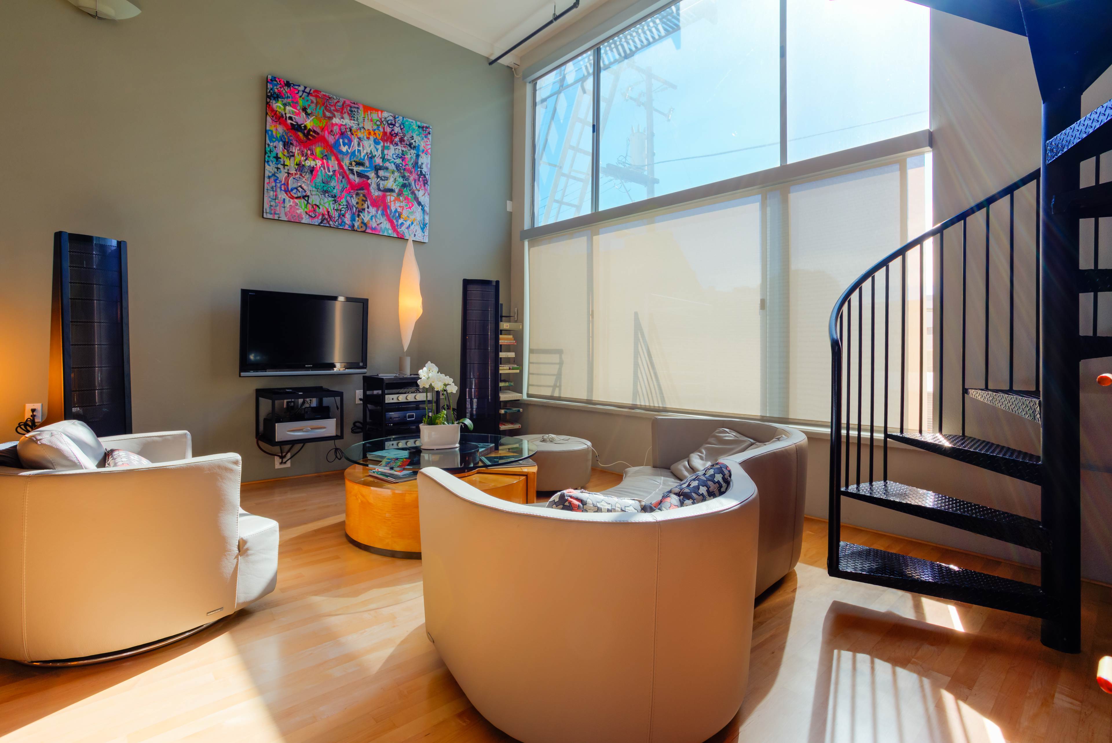 The living room features a spiral staircase, large windows with natural light, modern furniture, and a colorful artwork on the wall.