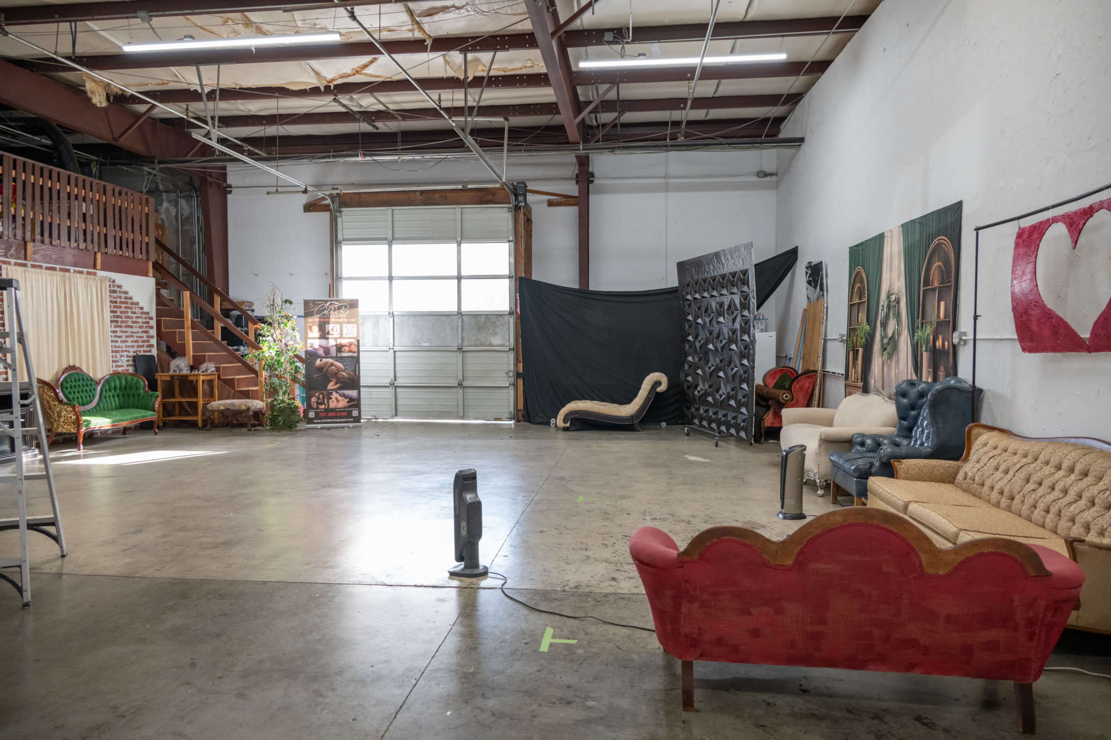 The image shows a spacious industrial studio with various couches arranged around a large open area, a staircase in the background, and different decorative backdrops leaning against the walls.