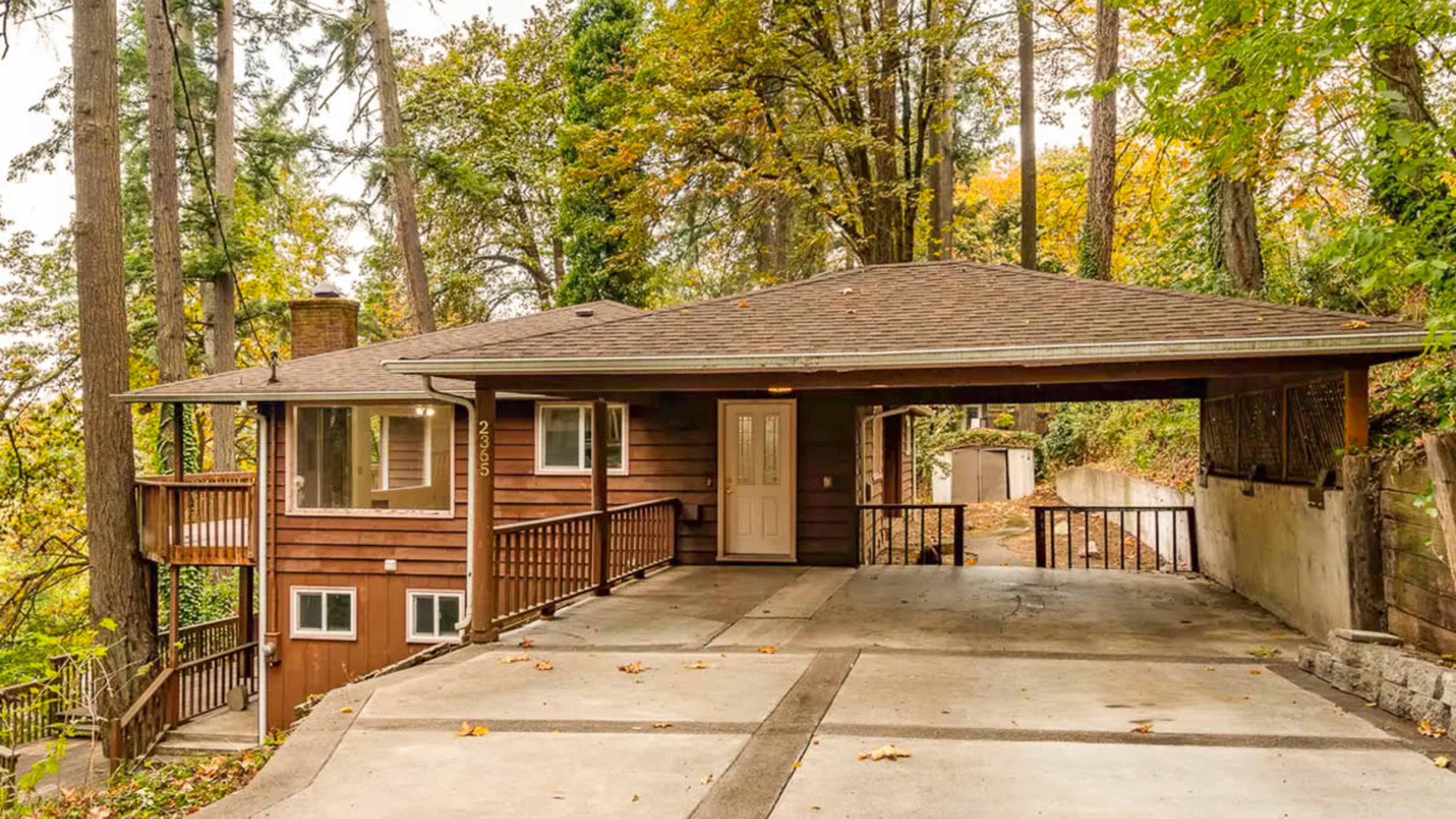 A brown wooden house with a large carport is surrounded by trees and a concrete driveway.