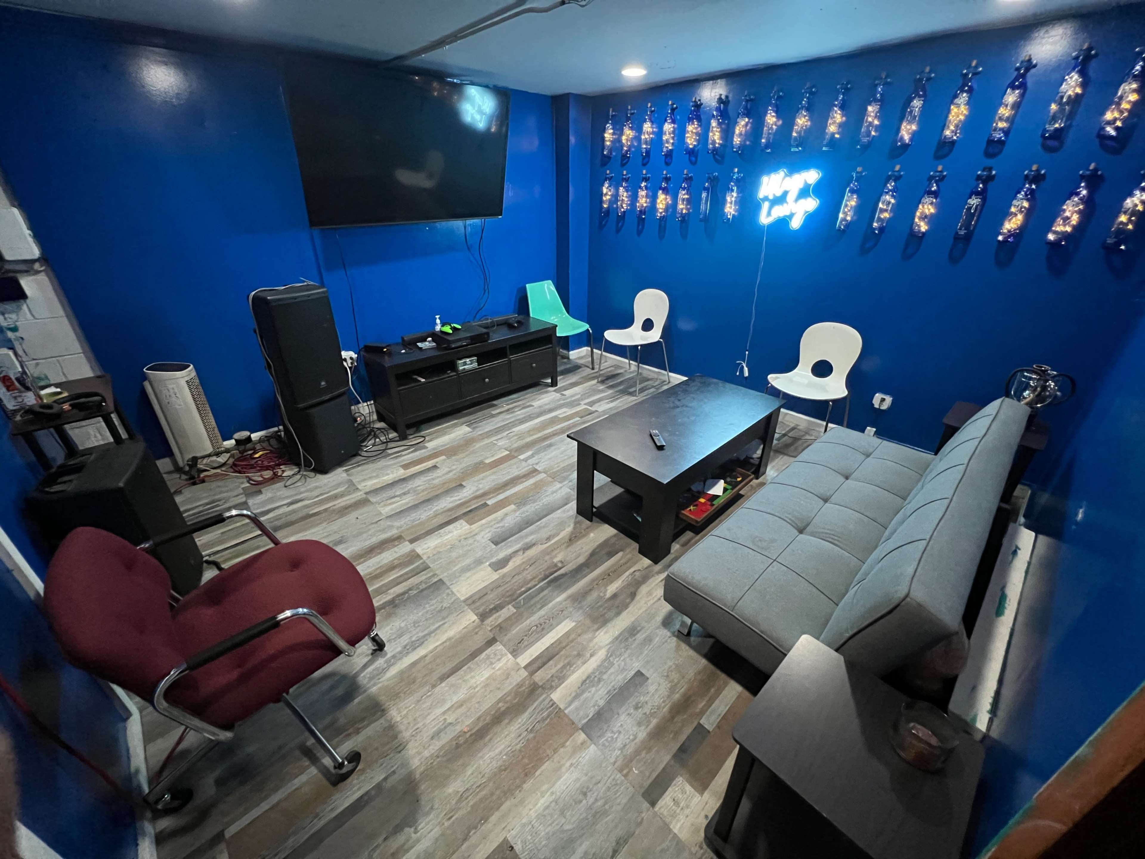 The room features a blue-painted wall with a large TV, a gray couch, a black coffee table, two white chairs, and a red office chair, all situated on a wood-patterned floor.