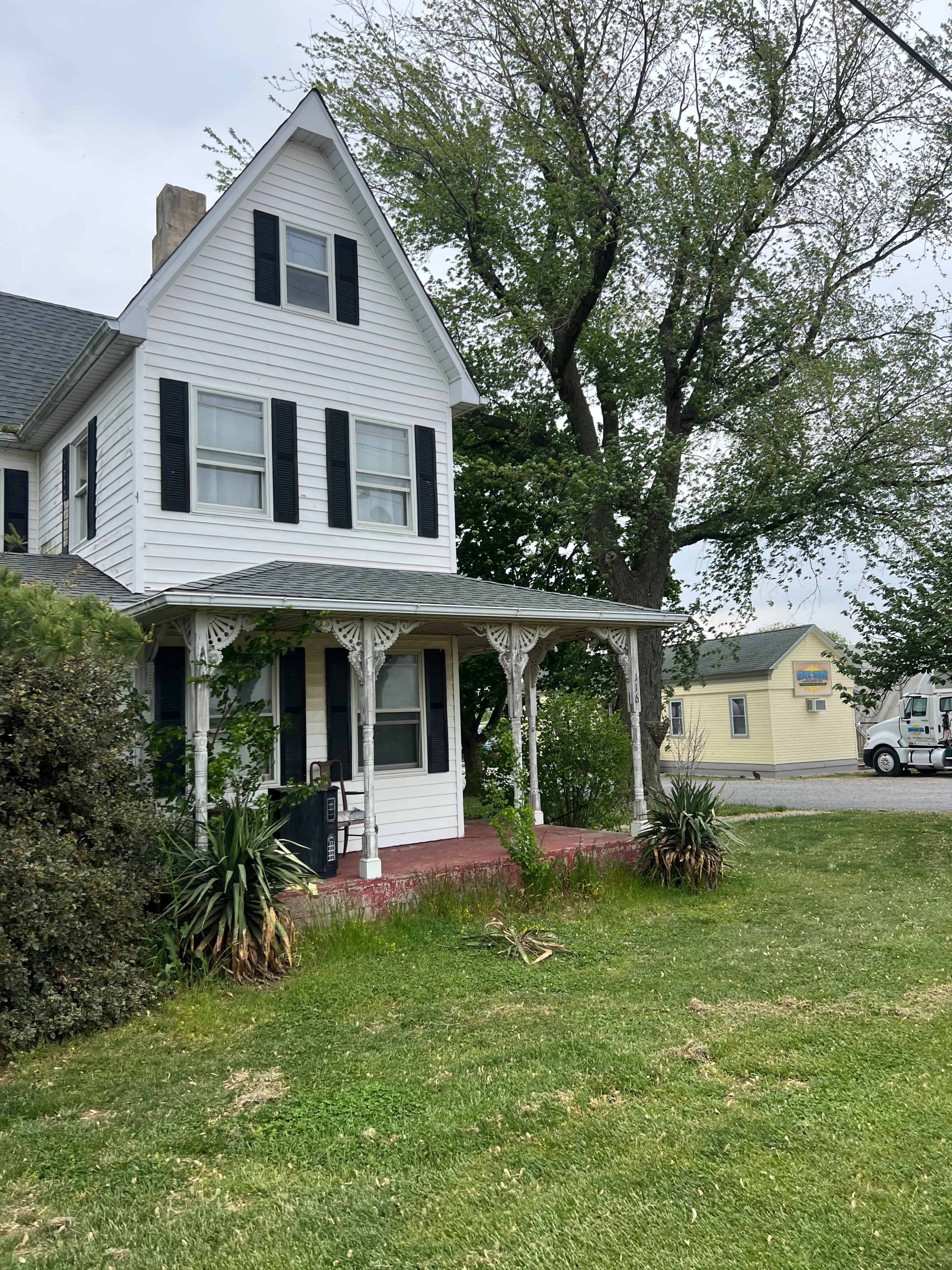 The image shows a two-story white house with black shutters and a porch adorned with decorative trim, surrounded by greenery and nearby structures.