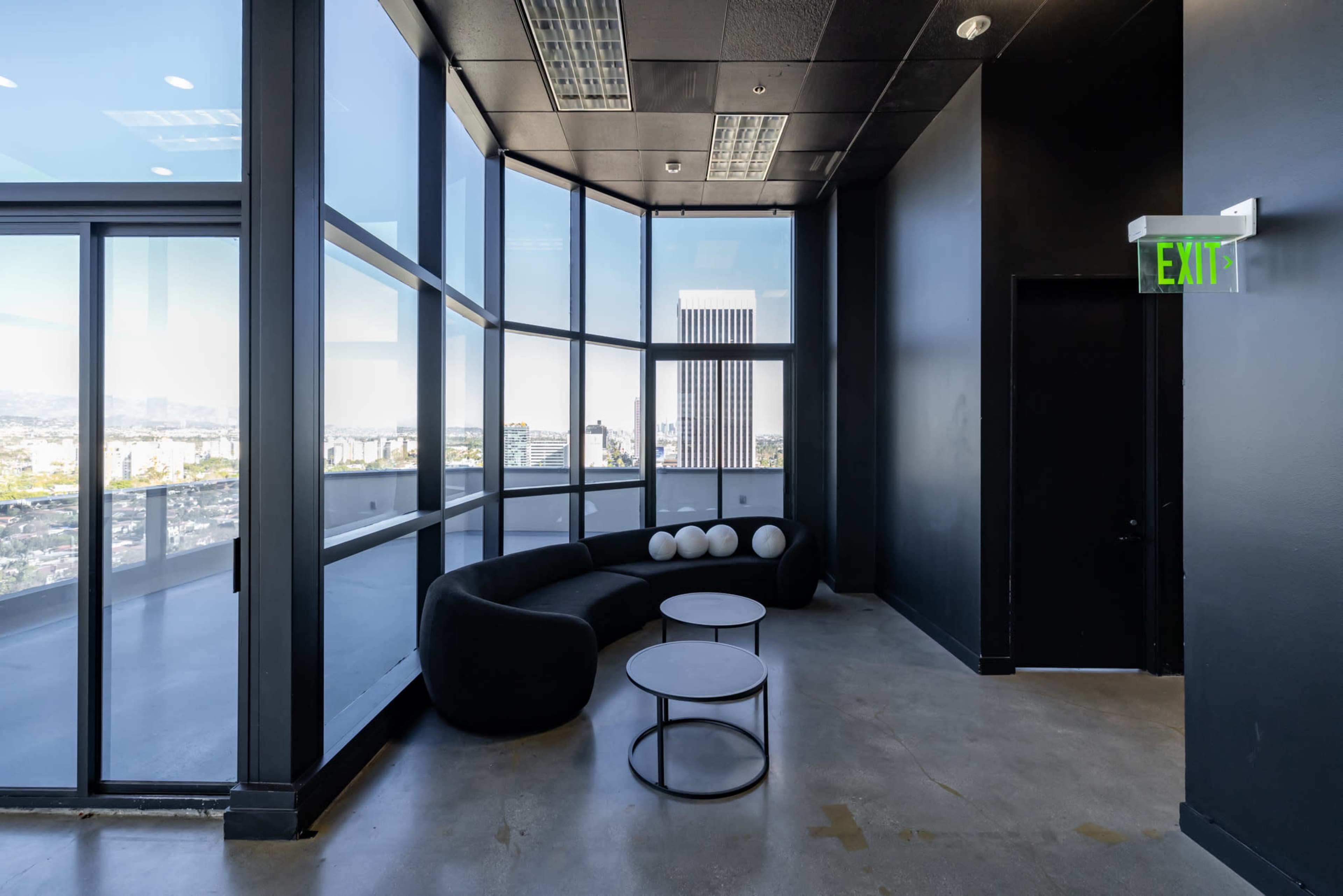 The image shows a modern lounge area with large glass windows, a curved black sofa, two white coffee tables, and a door marked "EXIT."