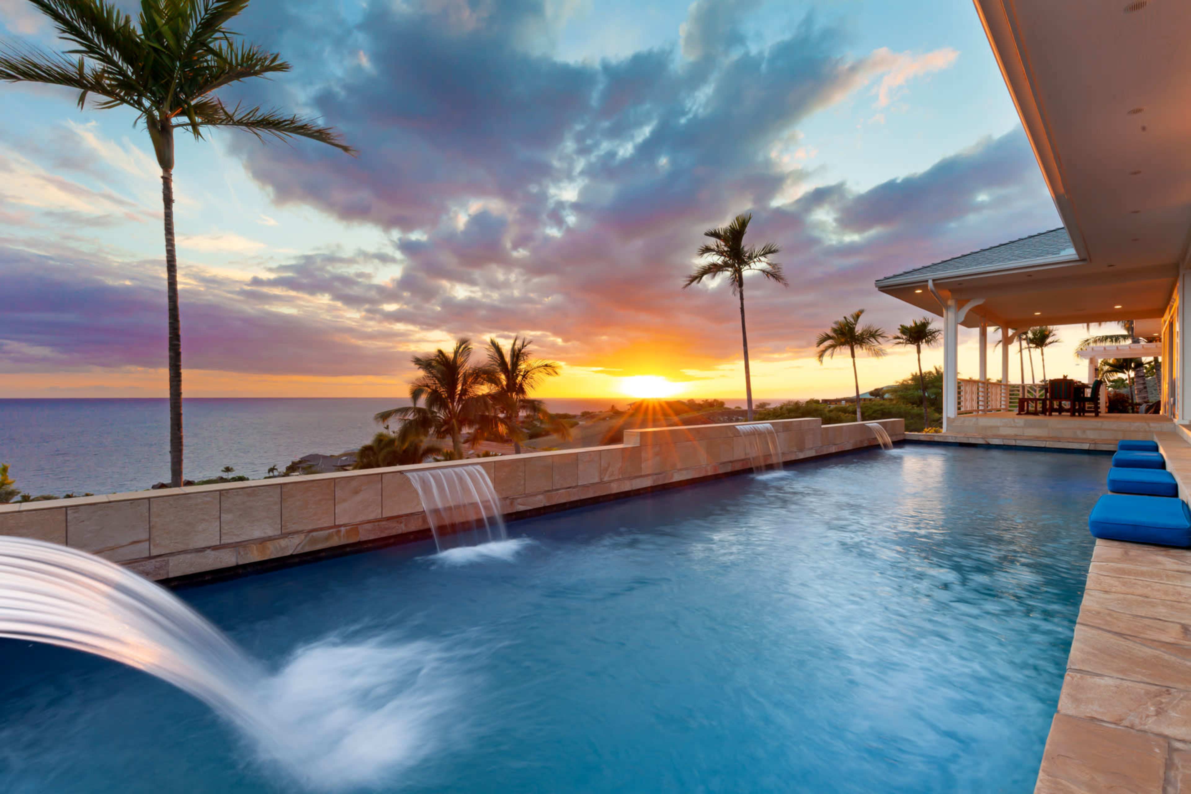 A luxurious infinity pool overlooks the ocean at sunset, framed by palm trees and a modern home.