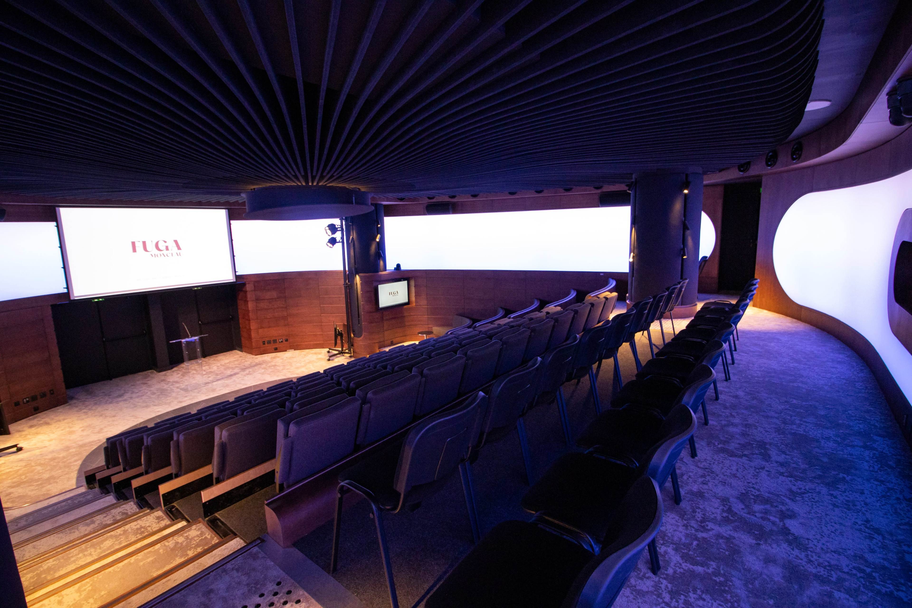 The image shows a modern auditorium with rows of seats facing a screen and softly lit walls.