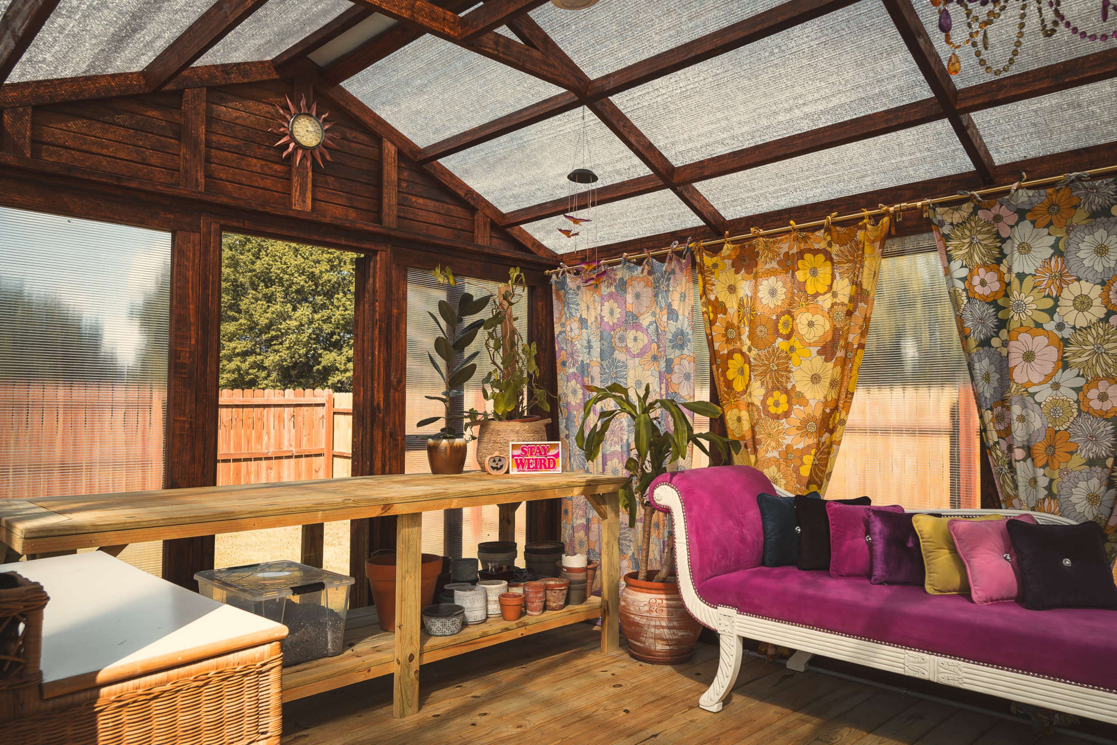 The image shows a wooden sunroom featuring a purple couch, colorful floral curtains, and a large wooden table with various plants and pots.