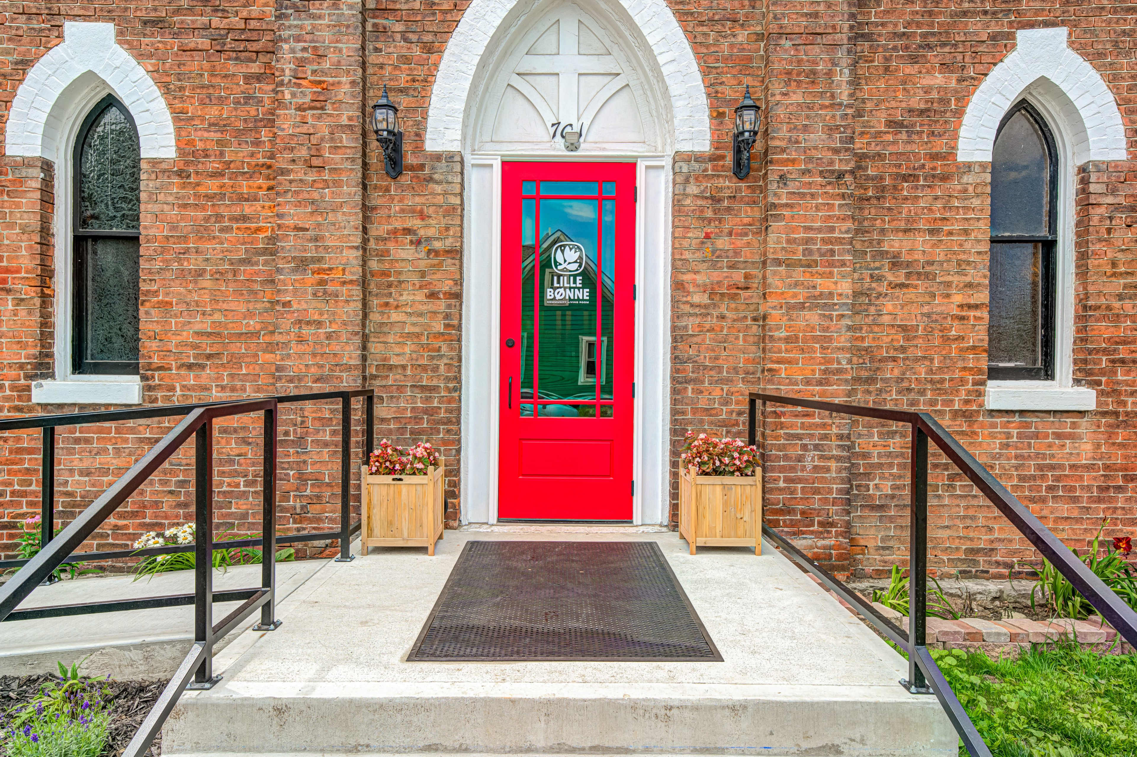 The image shows a red door with decorative glass, framed by brick walls and flanked by two planters, leading into a building.