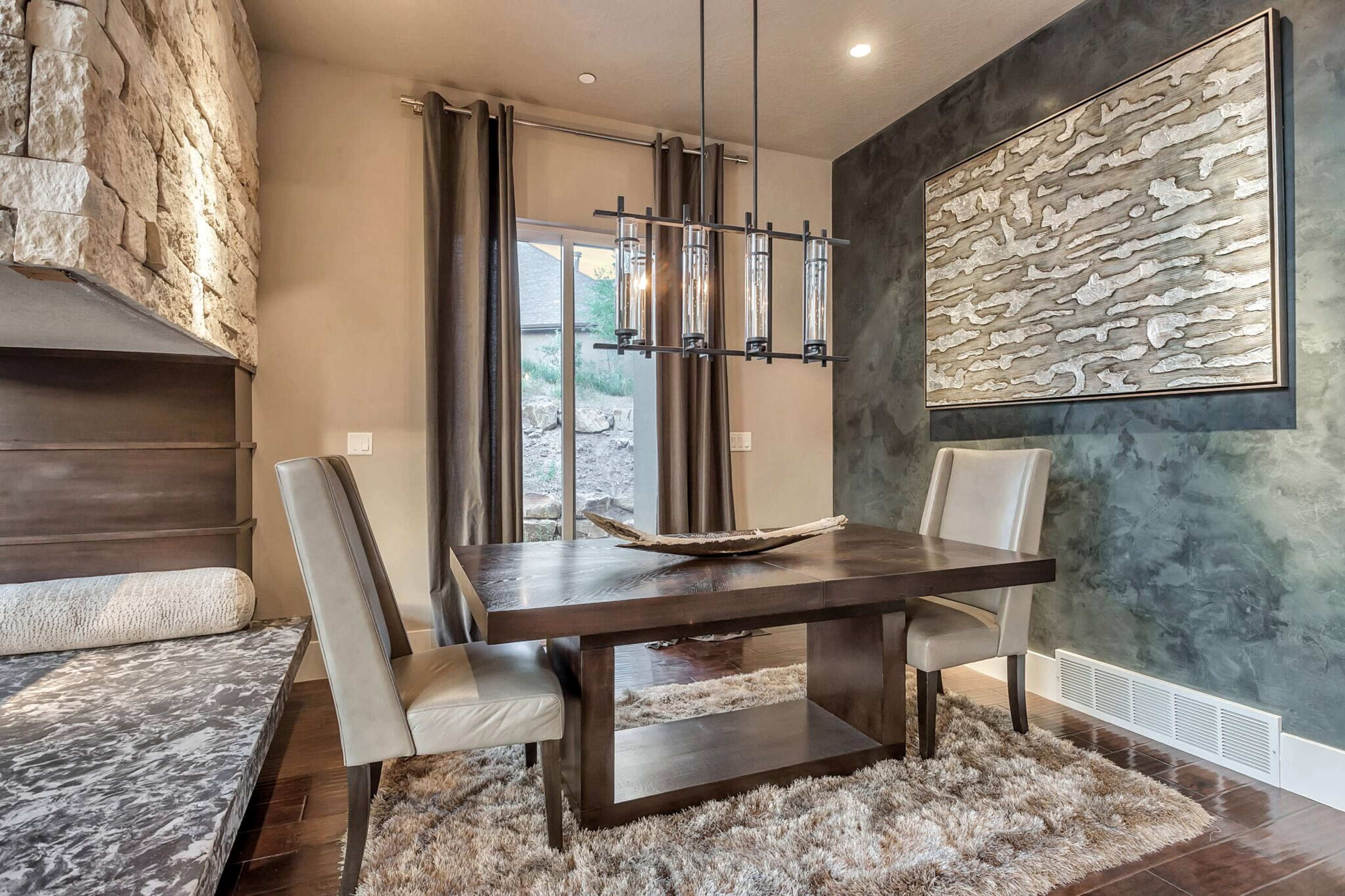 A modern dining room features a wooden table surrounded by two upholstered chairs, with a stone accent wall and a large artwork above the table.