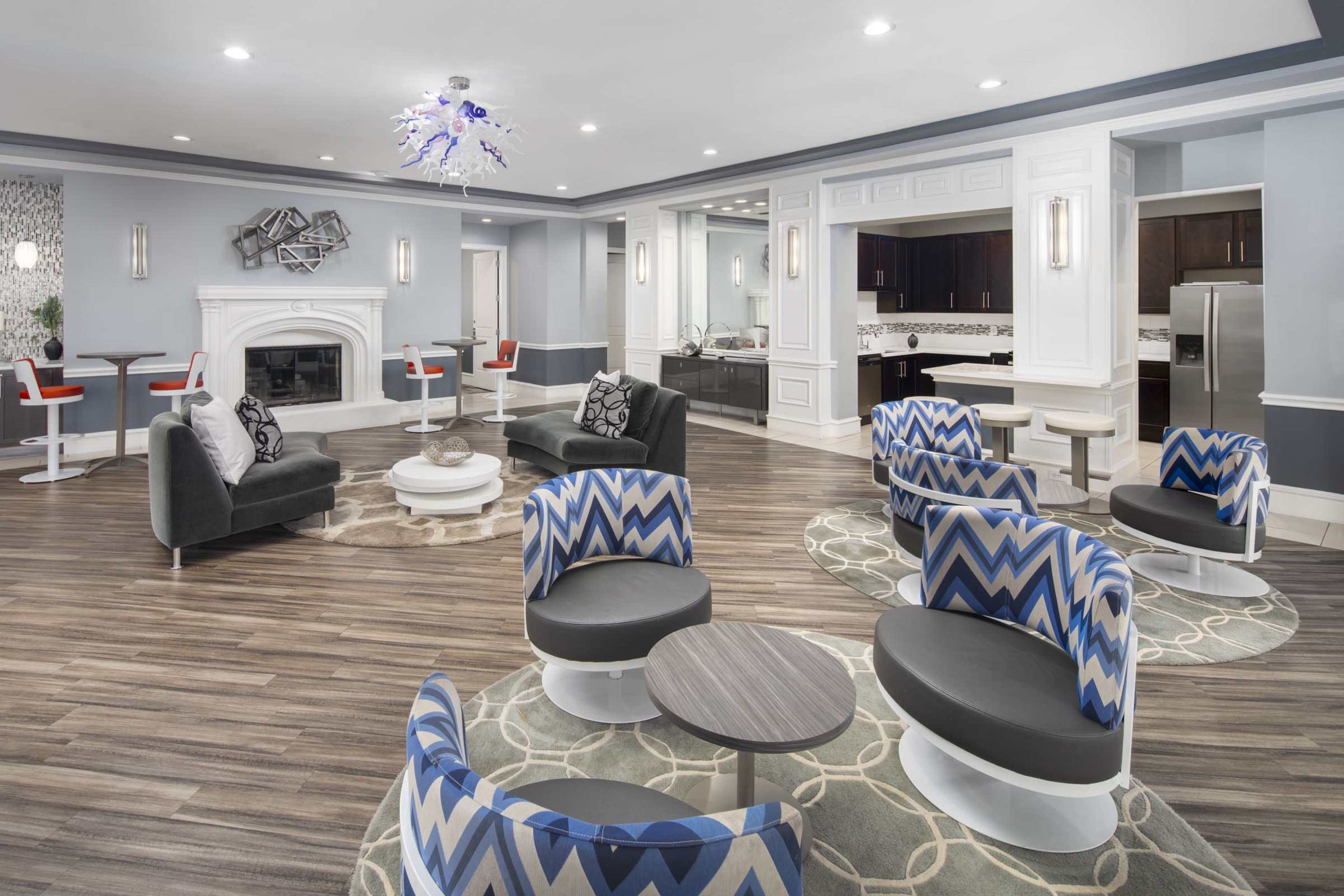 The image shows a modern lounge area featuring various seating arrangements, a fireplace, and a kitchenette in the background.