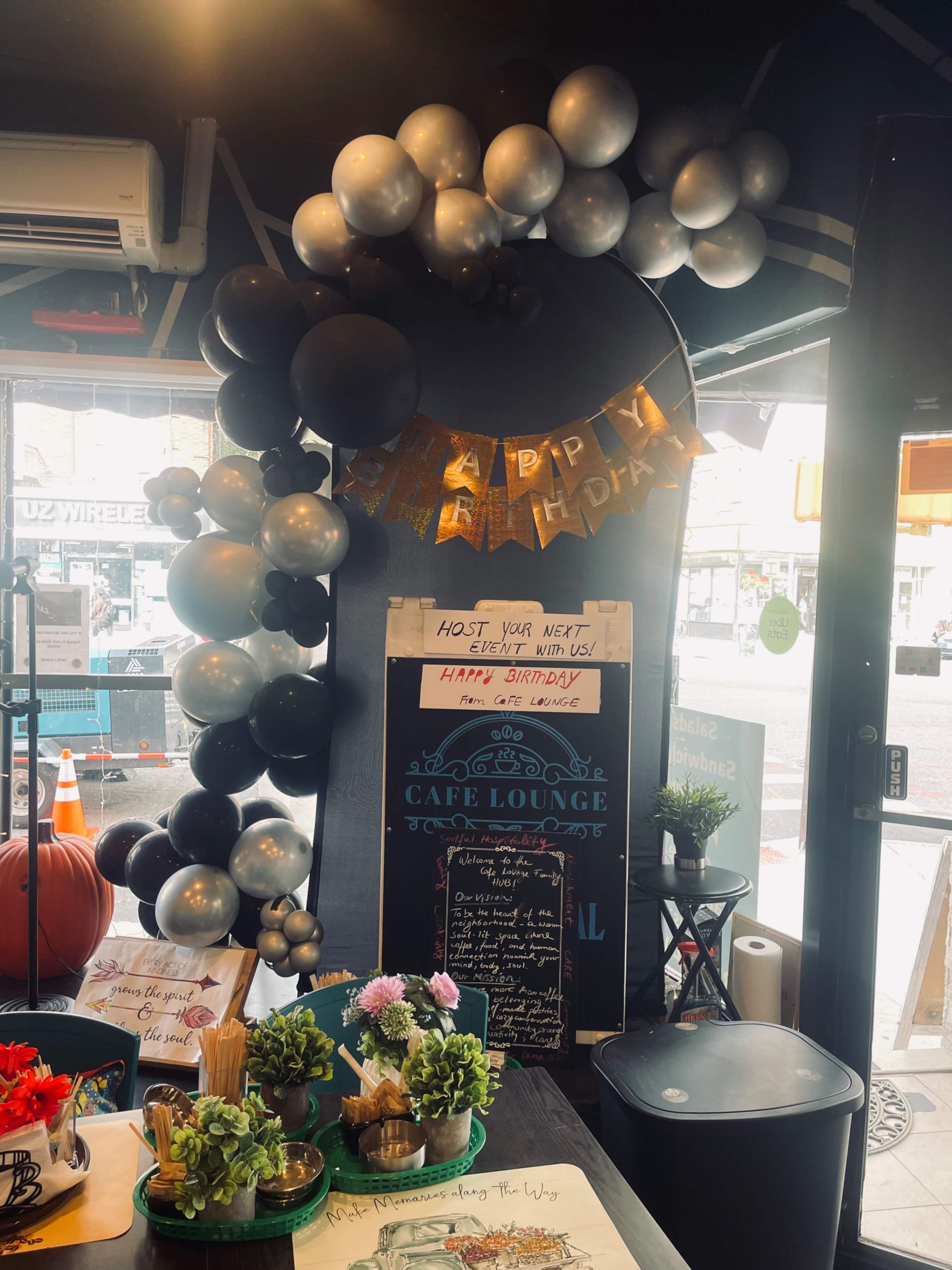 The image shows a festive birthday decoration featuring a balloon arch and a banner reading "HAPPY BIRTHDAY" inside a café lounge.
