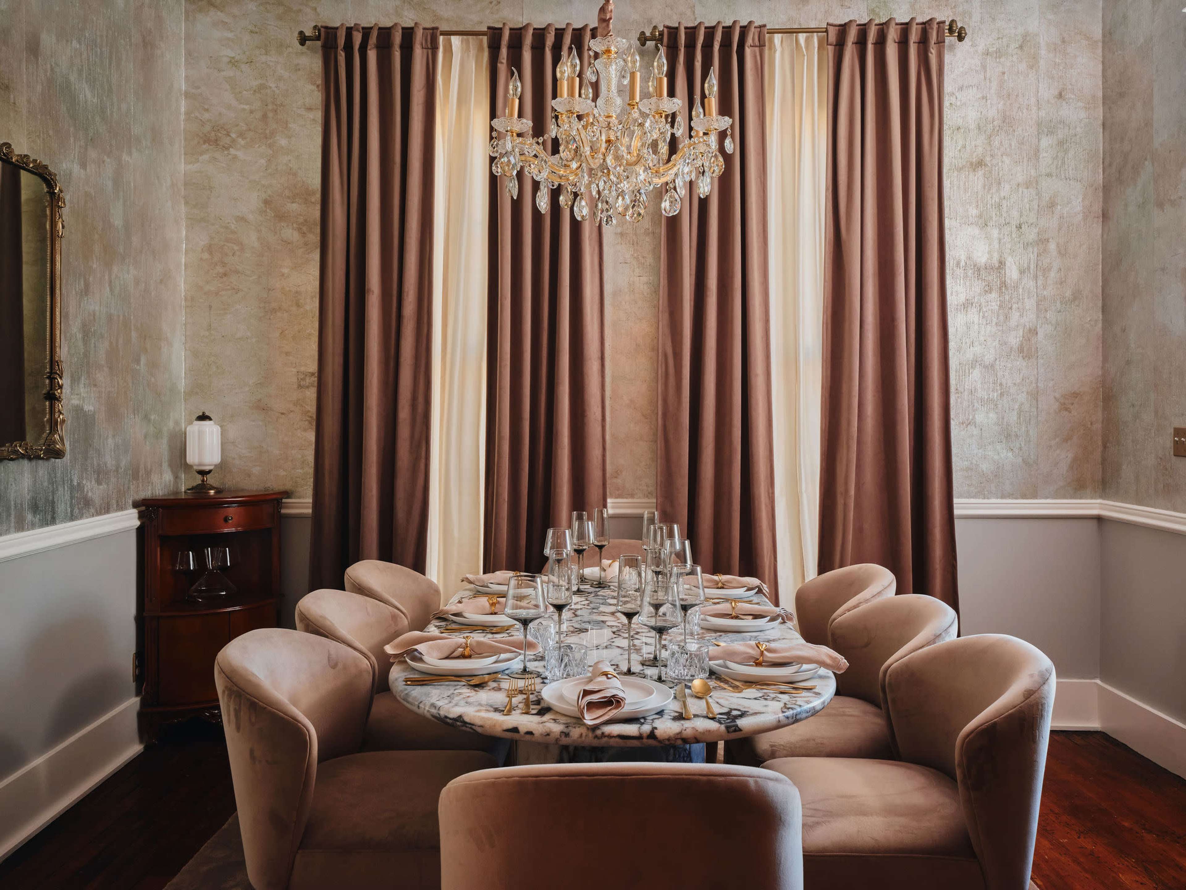 The image shows a dining room featuring a round marble table set for a meal, surrounded by eight upholstered chairs and adorned with a crystal chandelier above.