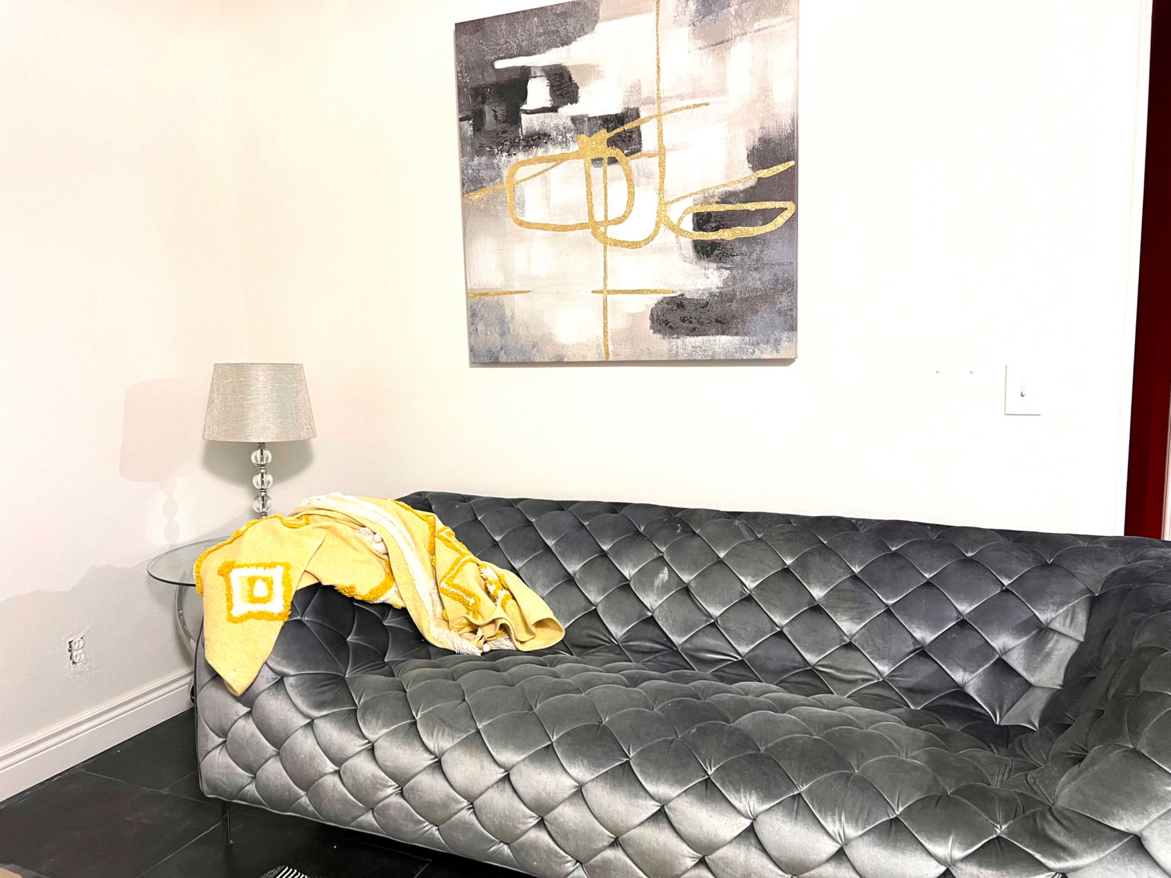 A grey tufted couch with a yellow and white blanket draped over it is positioned next to a lamp and a geometric artwork on the wall.