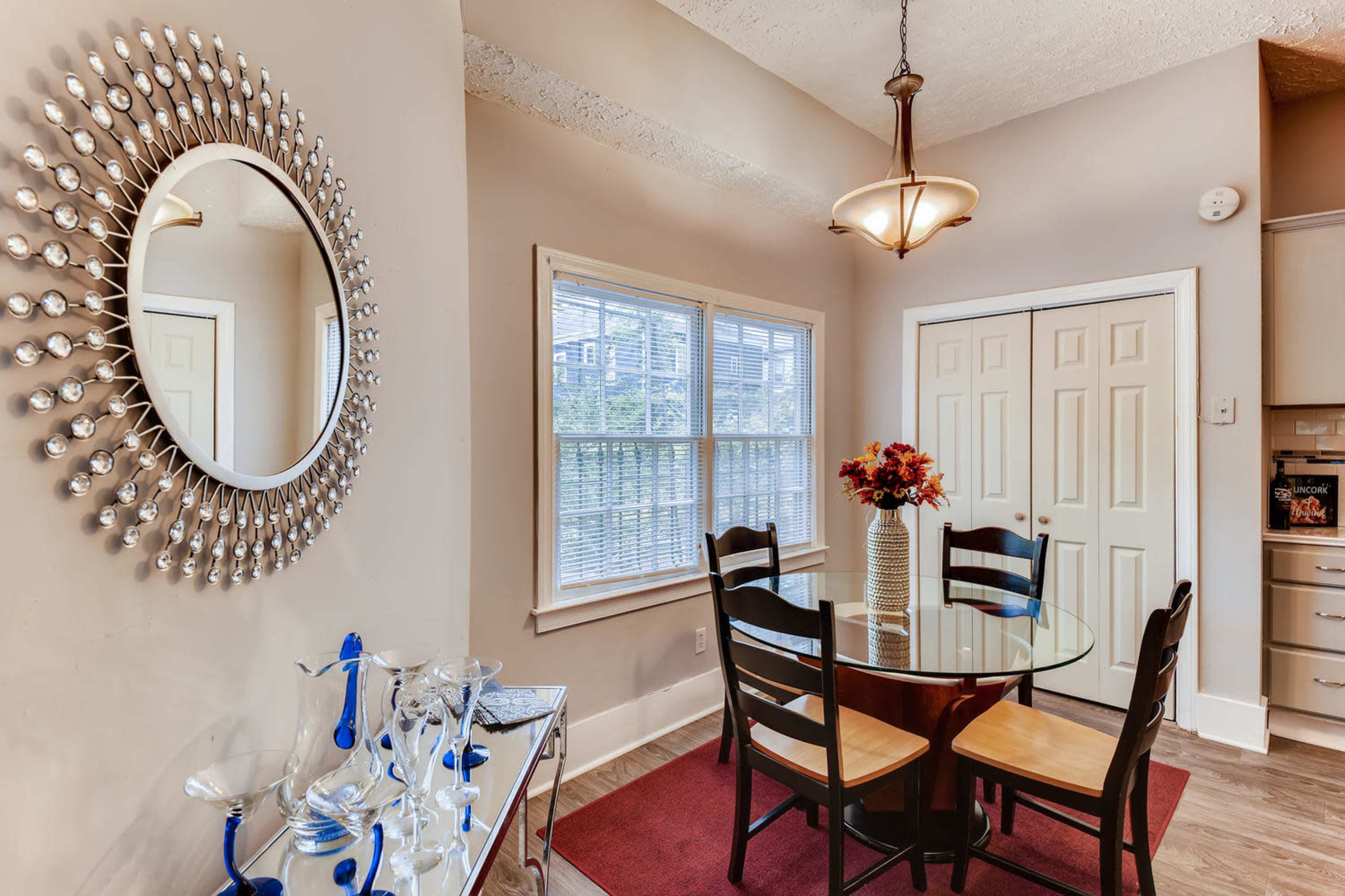A dining area features a round glass table surrounded by four black chairs, a vase of flowers in the center, and a large mirror on the wall.