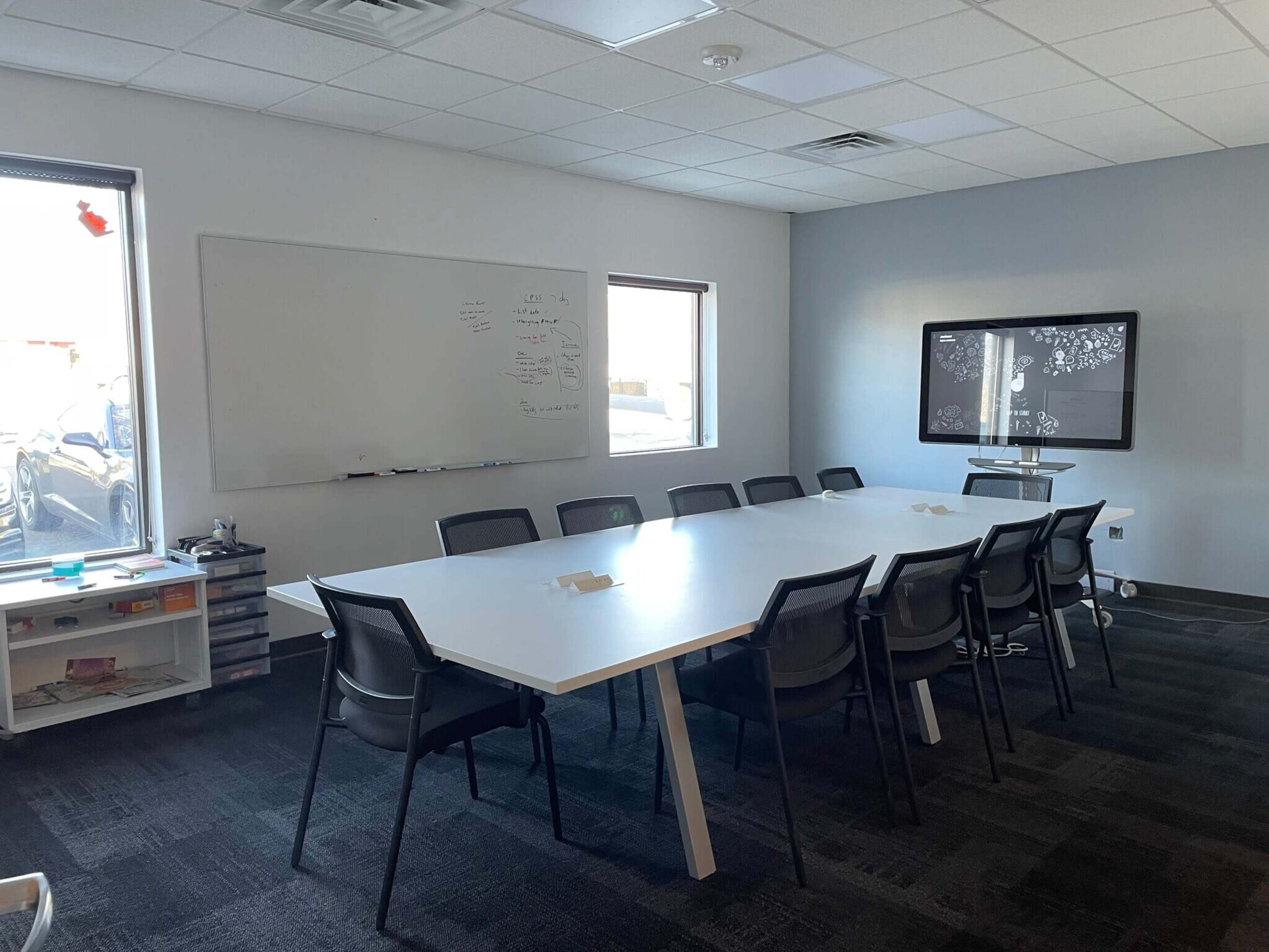 The image shows a large conference room with a long white table surrounded by black chairs, a whiteboard on one wall, and a screen on another wall.