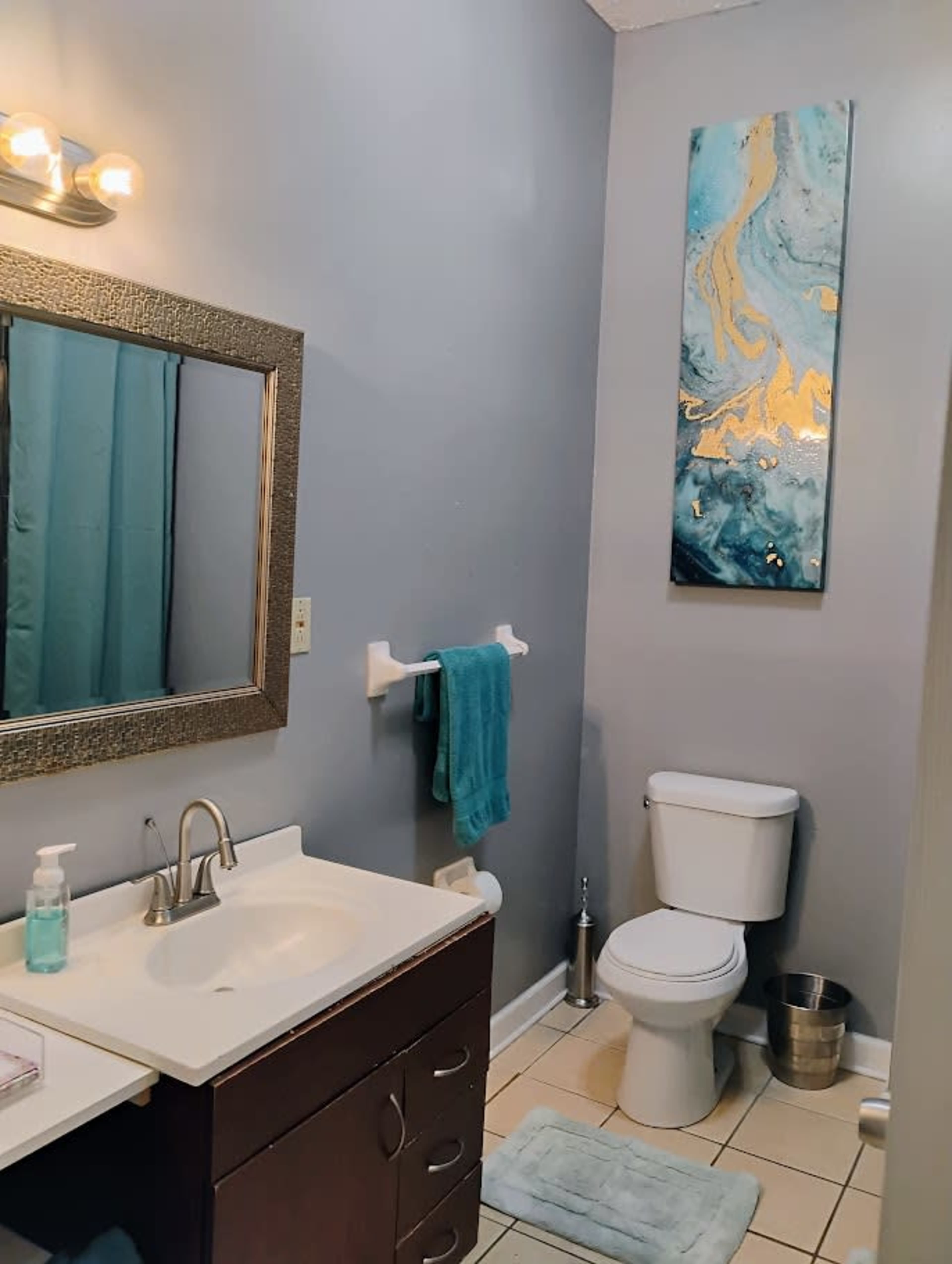 The image shows a small bathroom with a sink, mirror, and toilet, featuring light blue walls and a piece of abstract art above the toilet.
