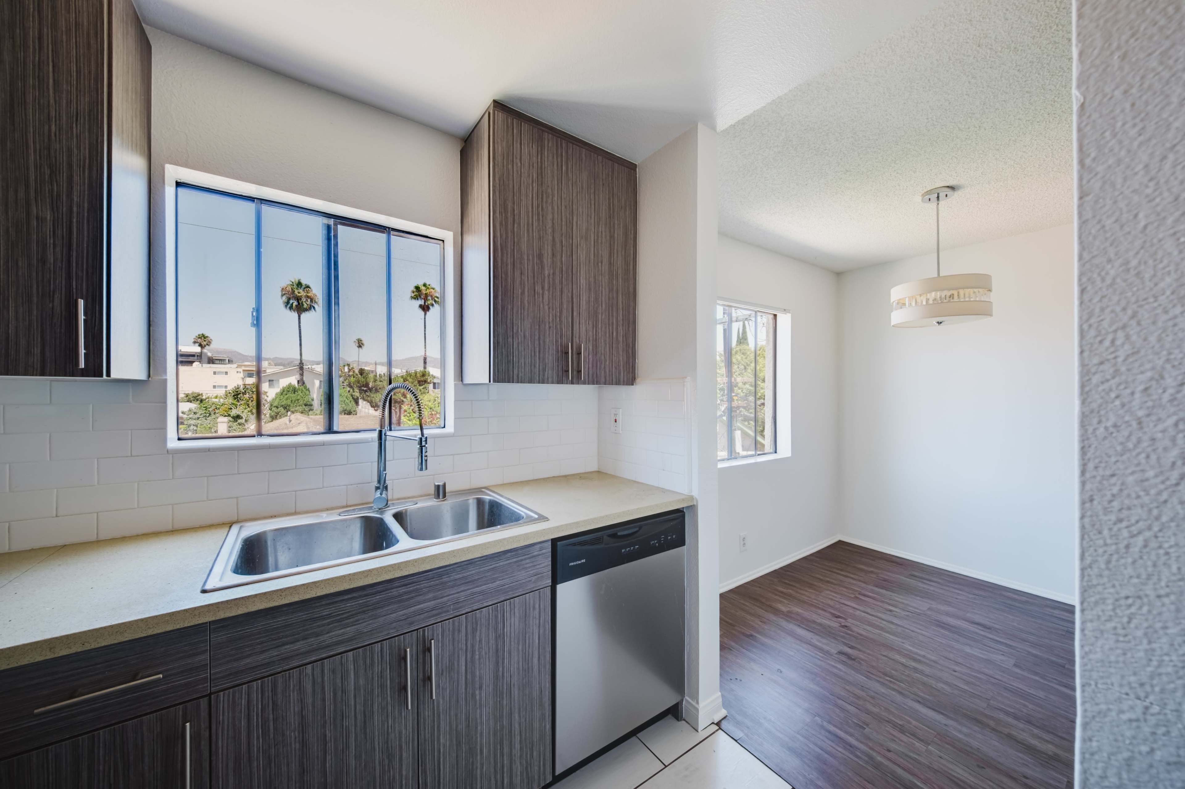 The image shows a modern kitchen with a double sink and a dishwasher, featuring dark wood cabinets, and a bright adjoining room with a circular light fixture and windows.