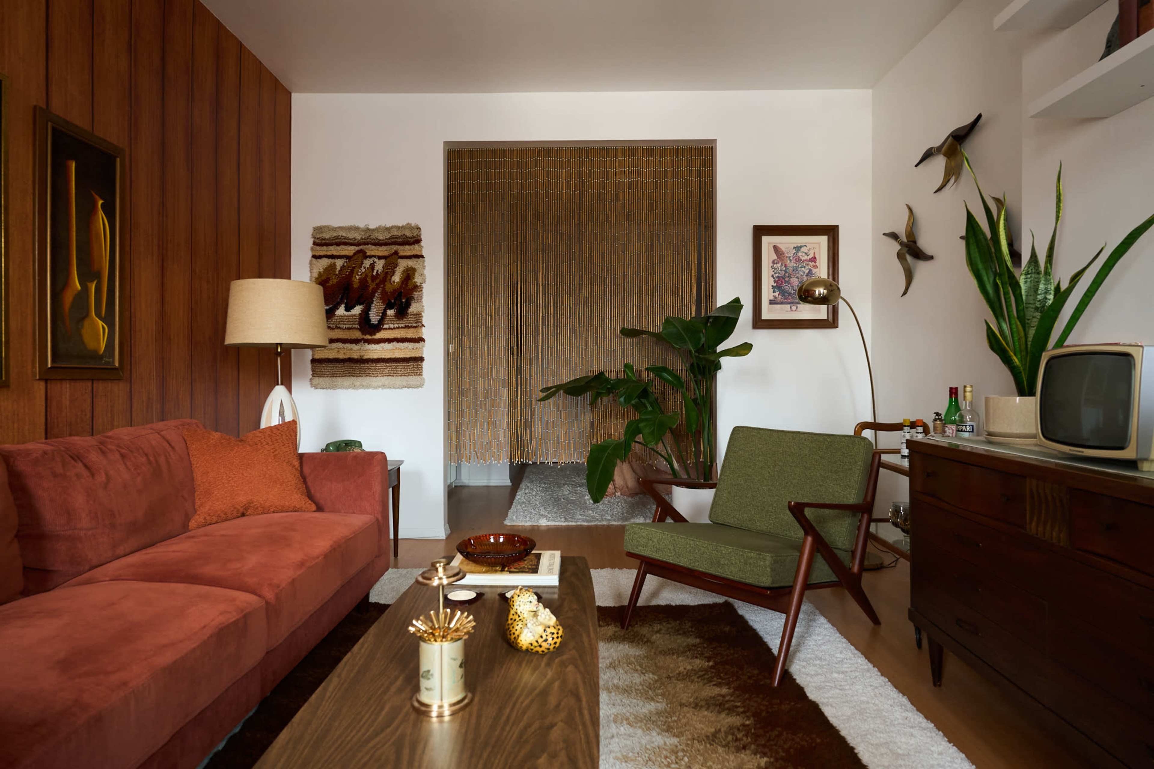 The image shows a mid-century modern living room with a brown sofa, a wooden coffee table, and a decorative wall featuring a macrame curtain and indoor plants.