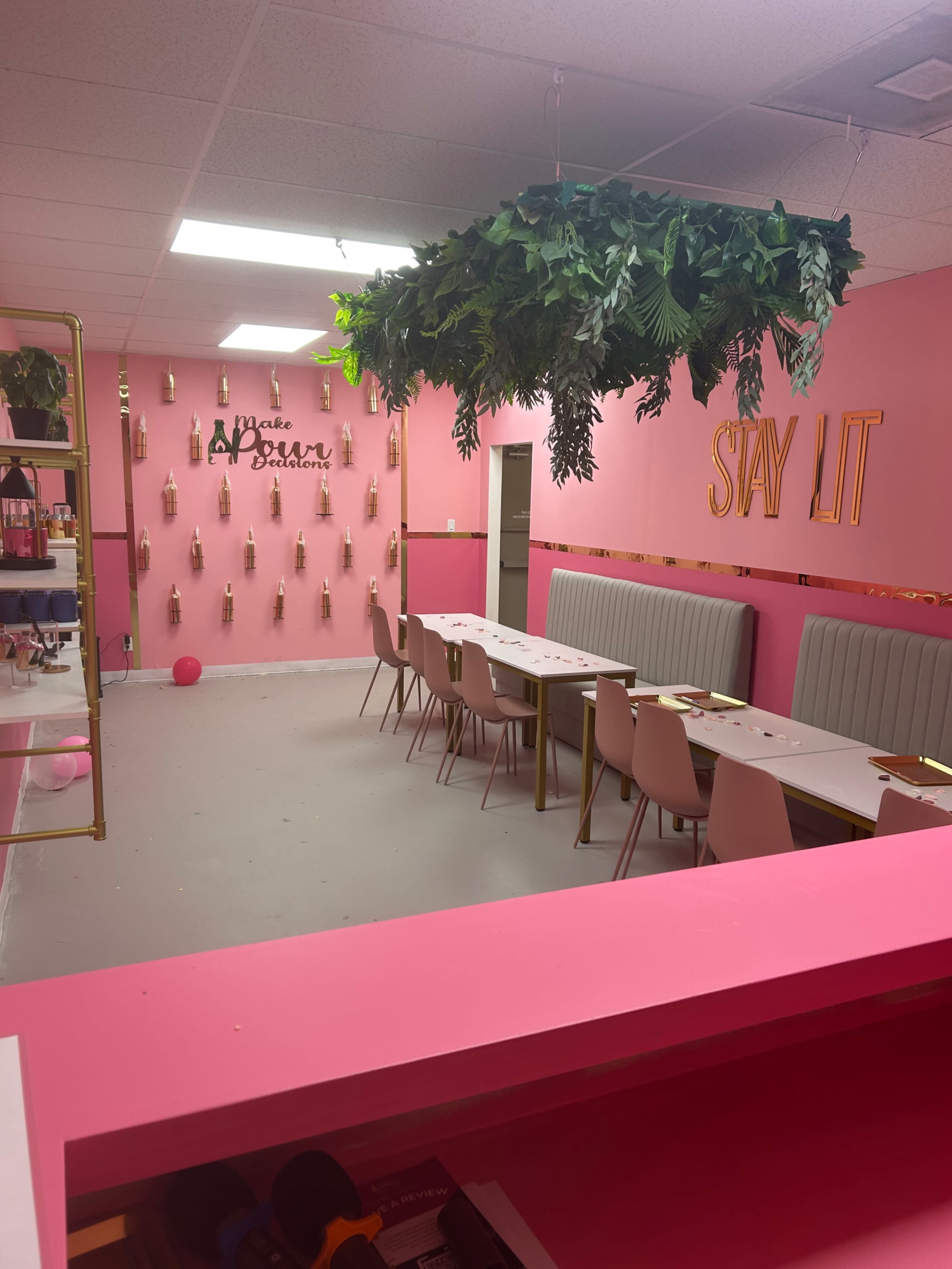 The image shows a pink-themed interior with tables and chairs arranged under a faux plant installation, featuring decorative wall signs and hanging bottles.