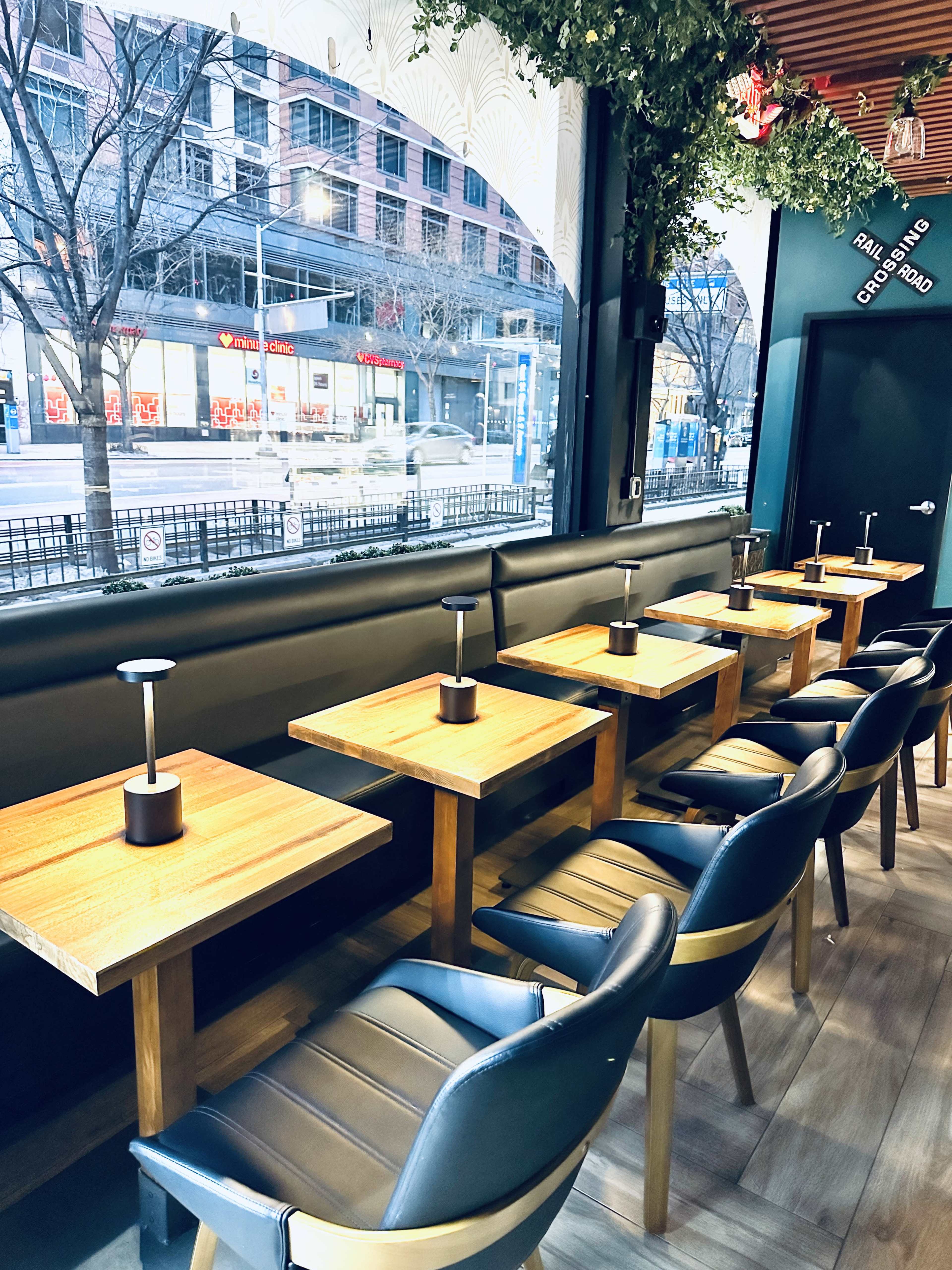 The image shows a row of wooden tables with black chairs beside large windows in a modern restaurant setting.