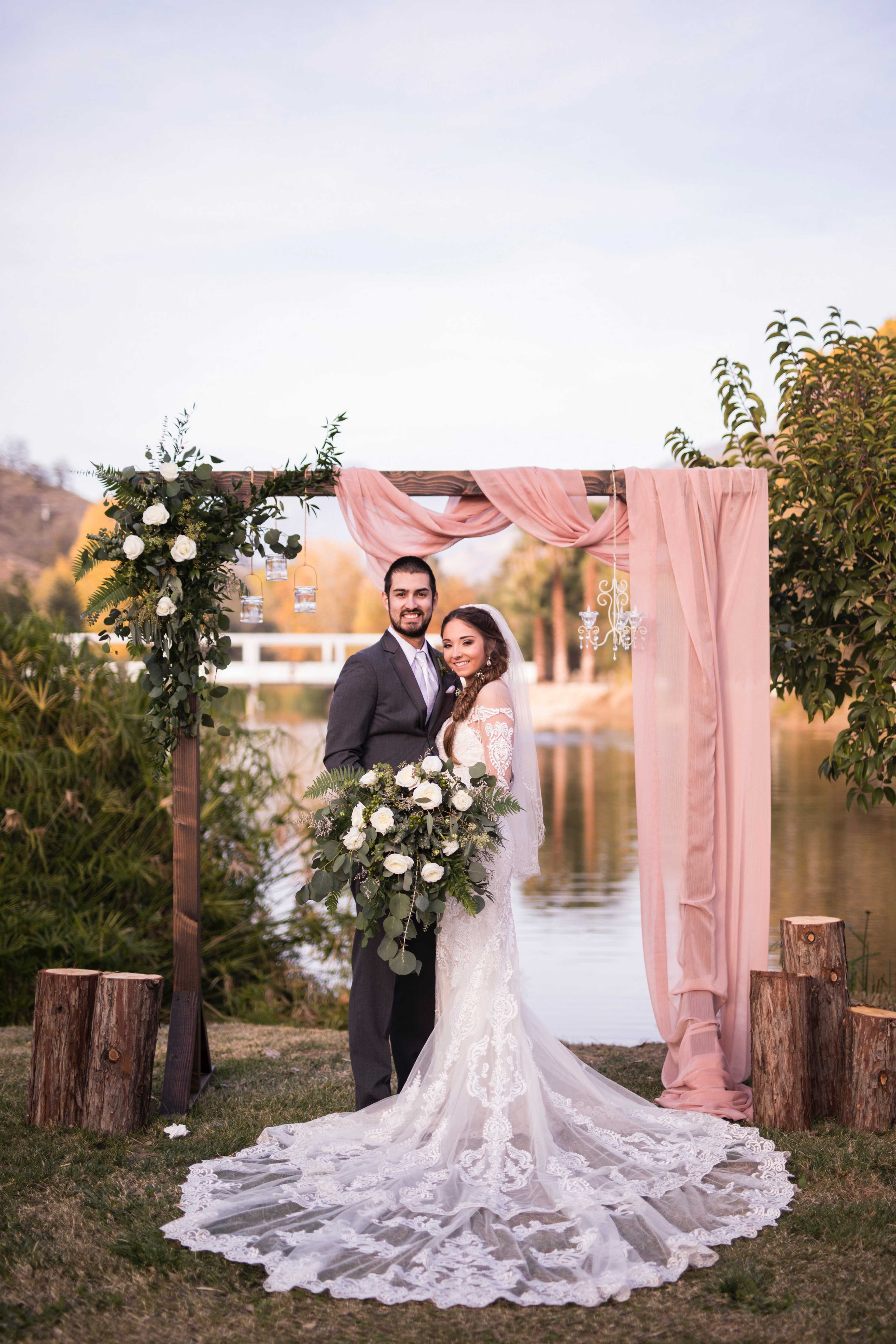 A bride and groom stand together under a floral arch beside a tranquil body of water.