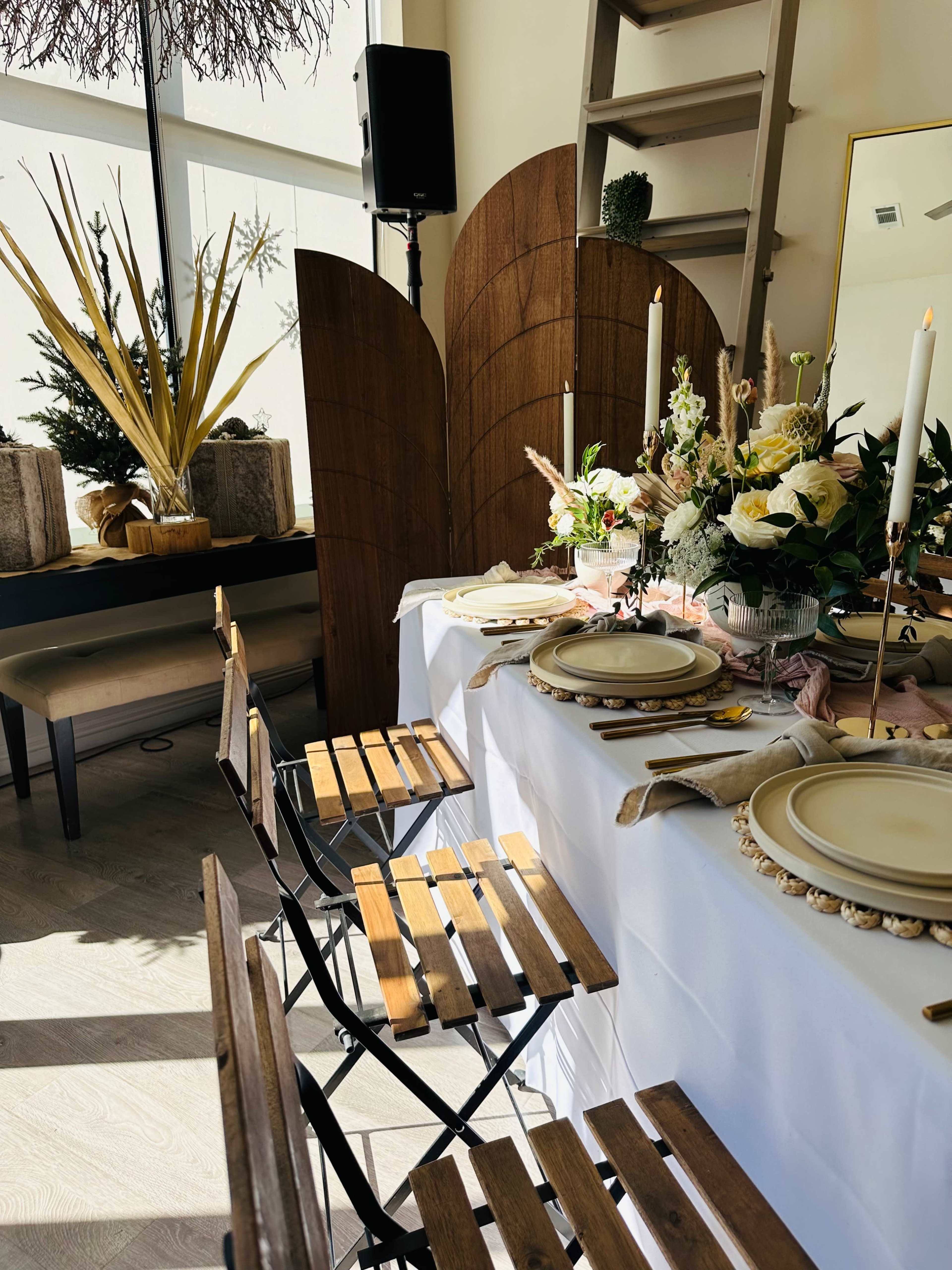 The image shows a dining table set with floral arrangements and candles, surrounded by wooden folding chairs in a bright indoor space.