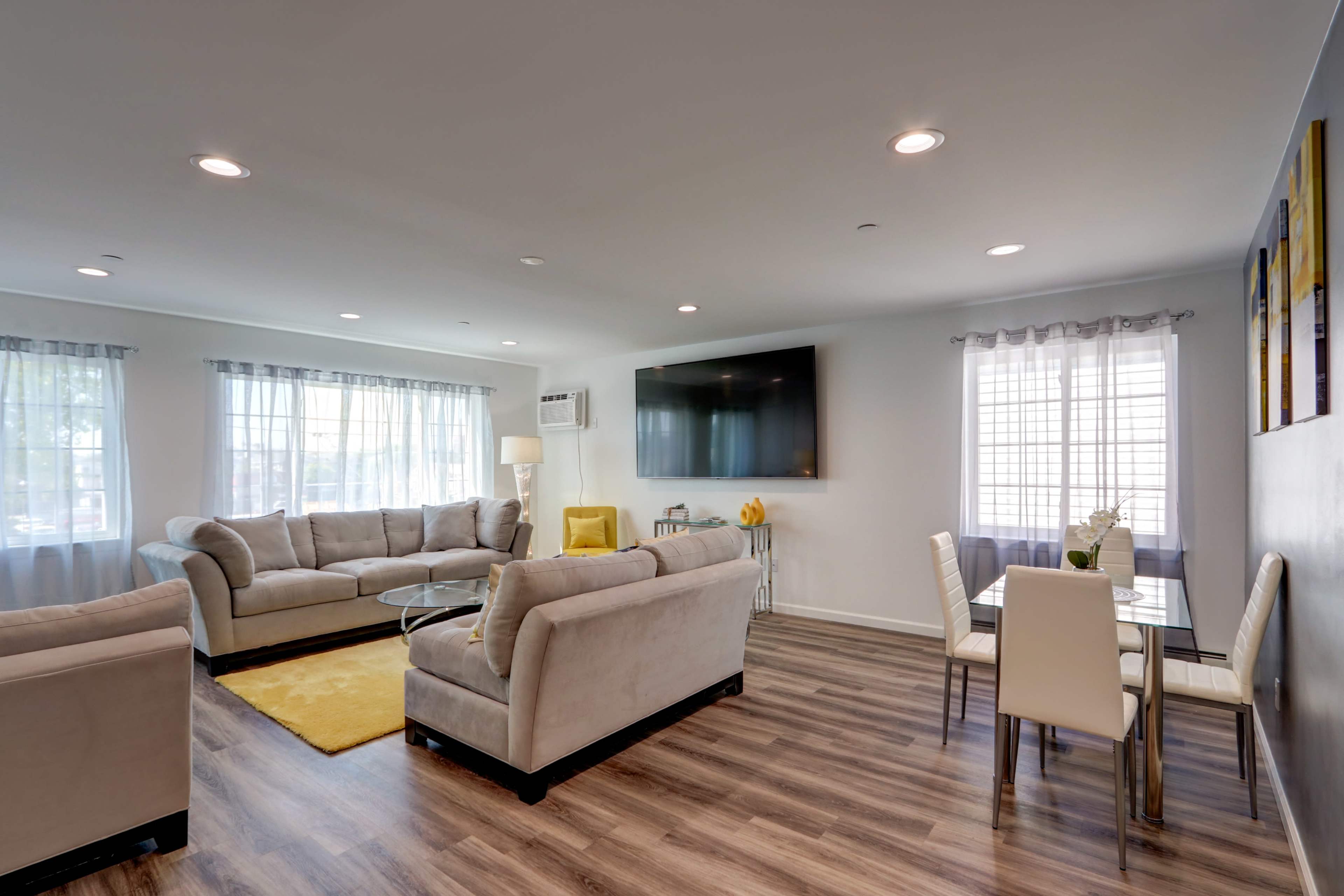 A modern living room with two gray sofas, a glass coffee table, a dining area with white chairs, and large windows allowing natural light.