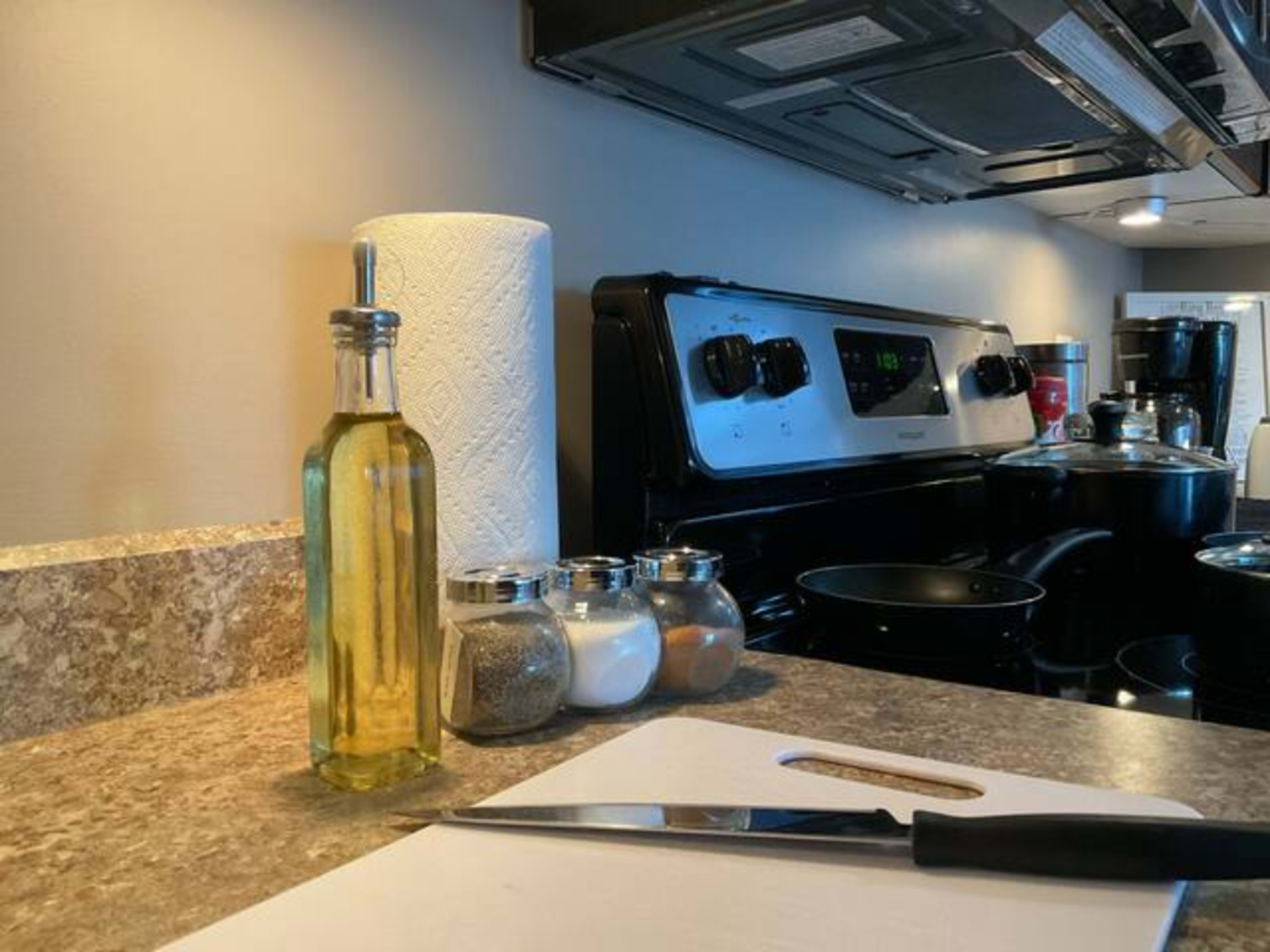 A countertop in a kitchen features a bottle of oil, jars of spices, a cutting board with a knife, and various cookware on the stove.