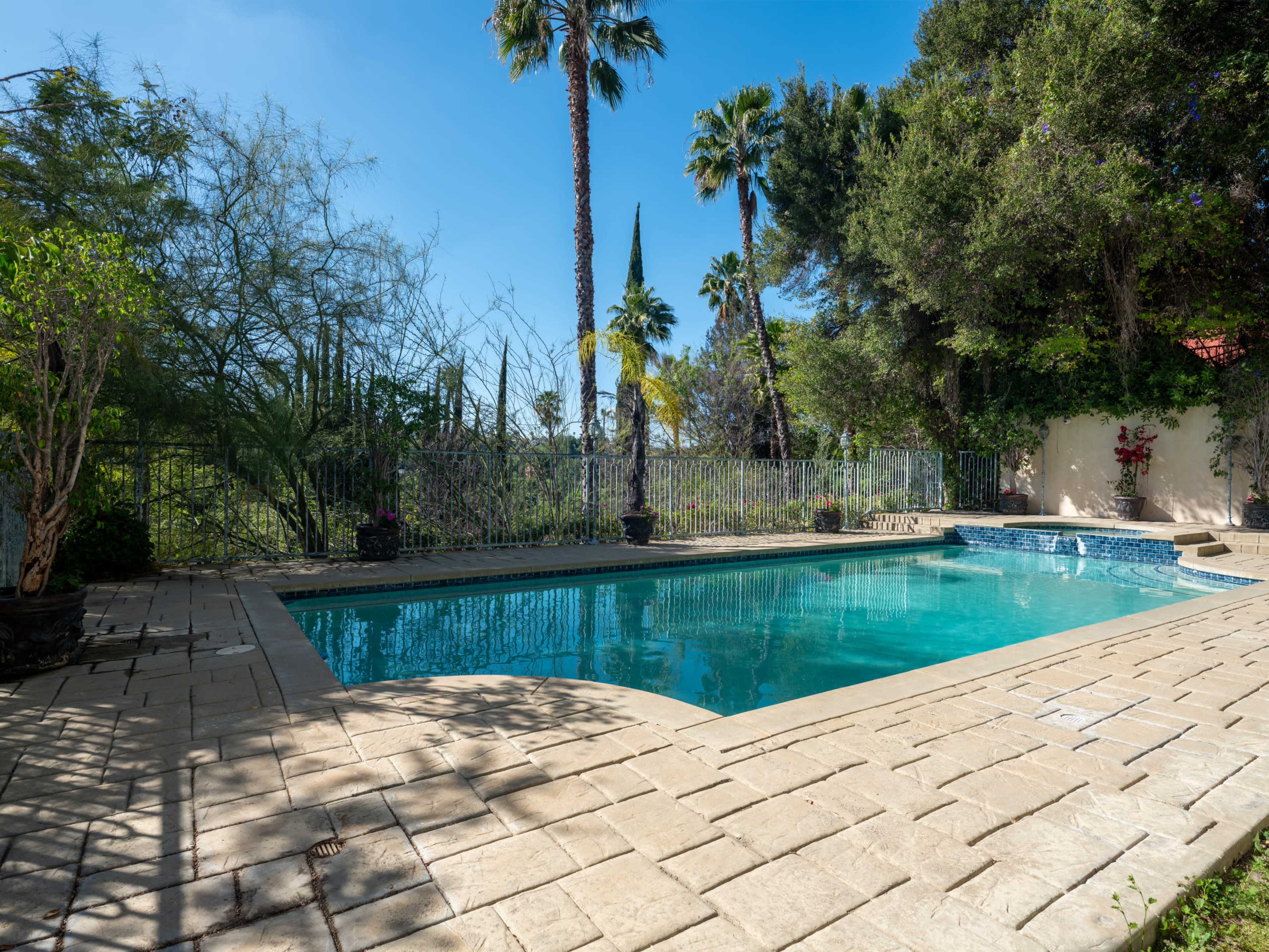 A clear swimming pool surrounded by tiled decking and palm trees under a blue sky.