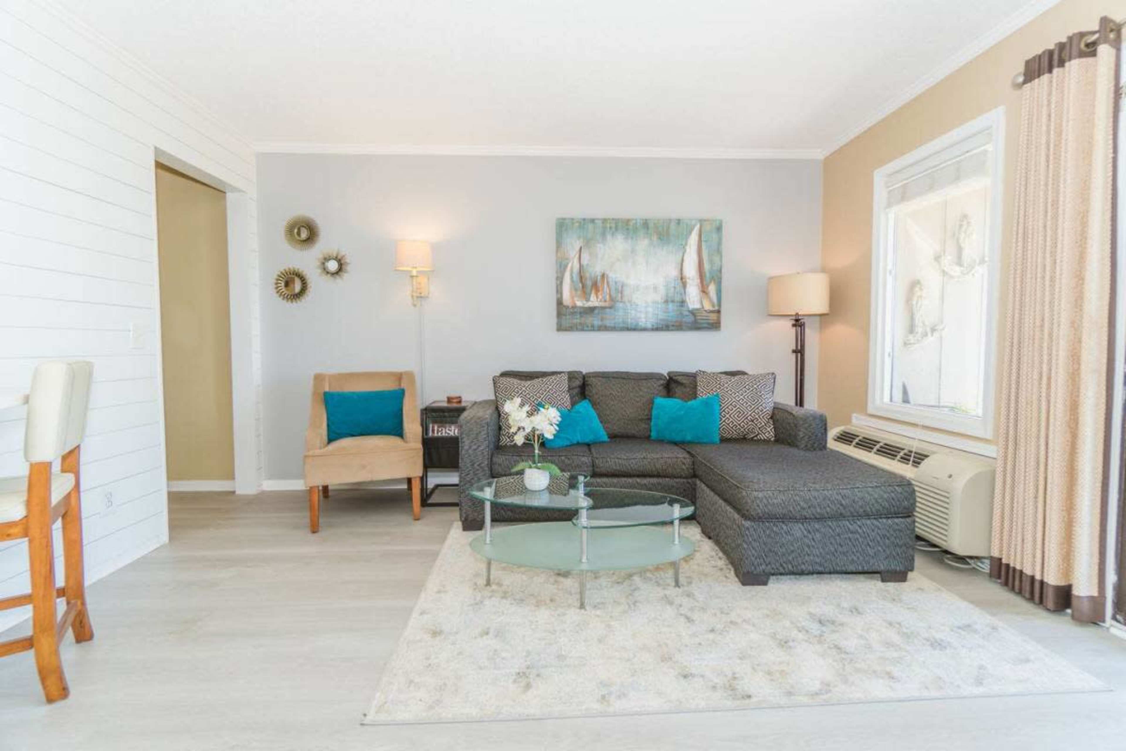 The image shows a modern living room with a gray sectional sofa, turquoise accent pillows, a glass coffee table, and light-colored wooden flooring.