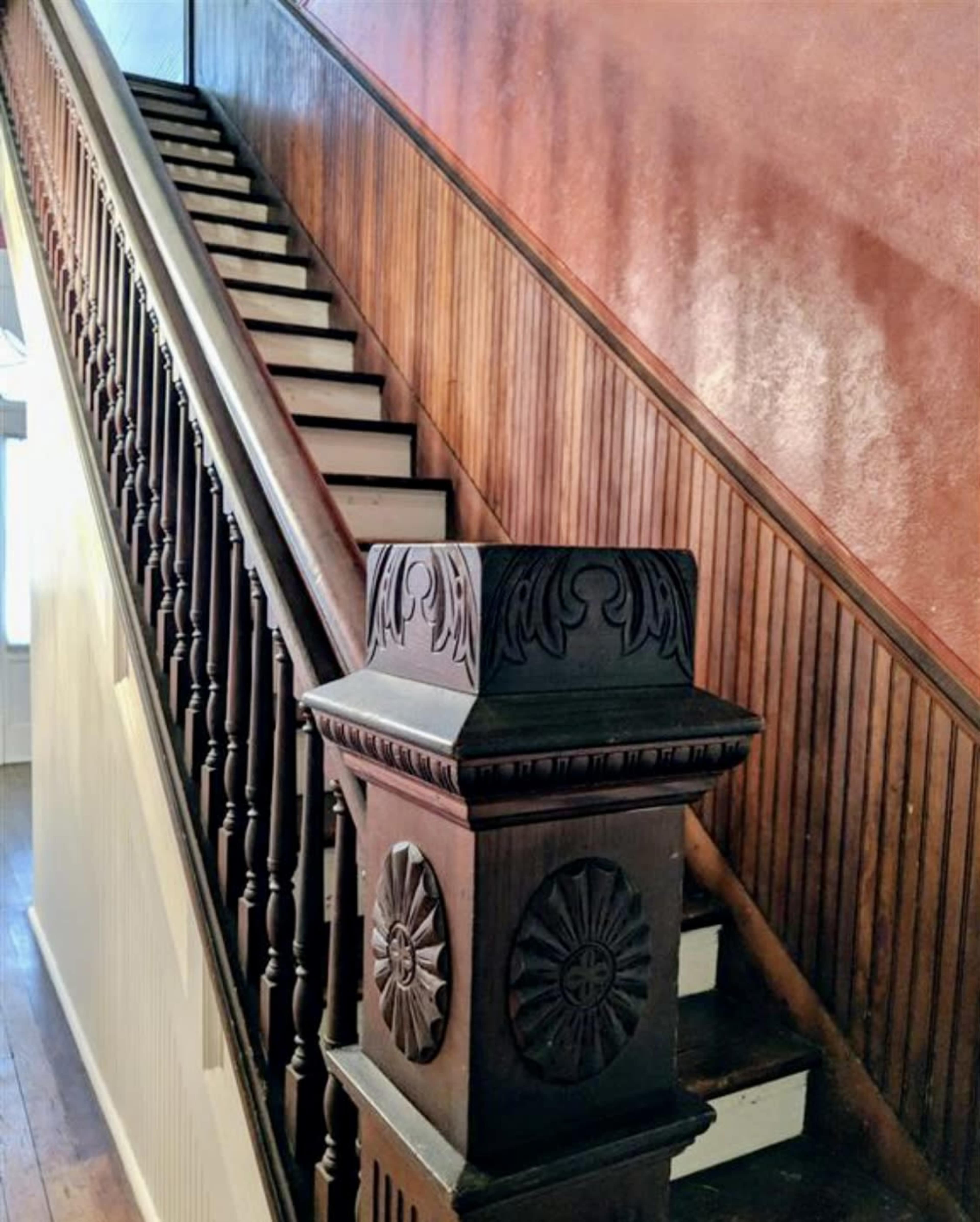 The image shows a wooden staircase with ornate railings leading upward against a paneled wall.