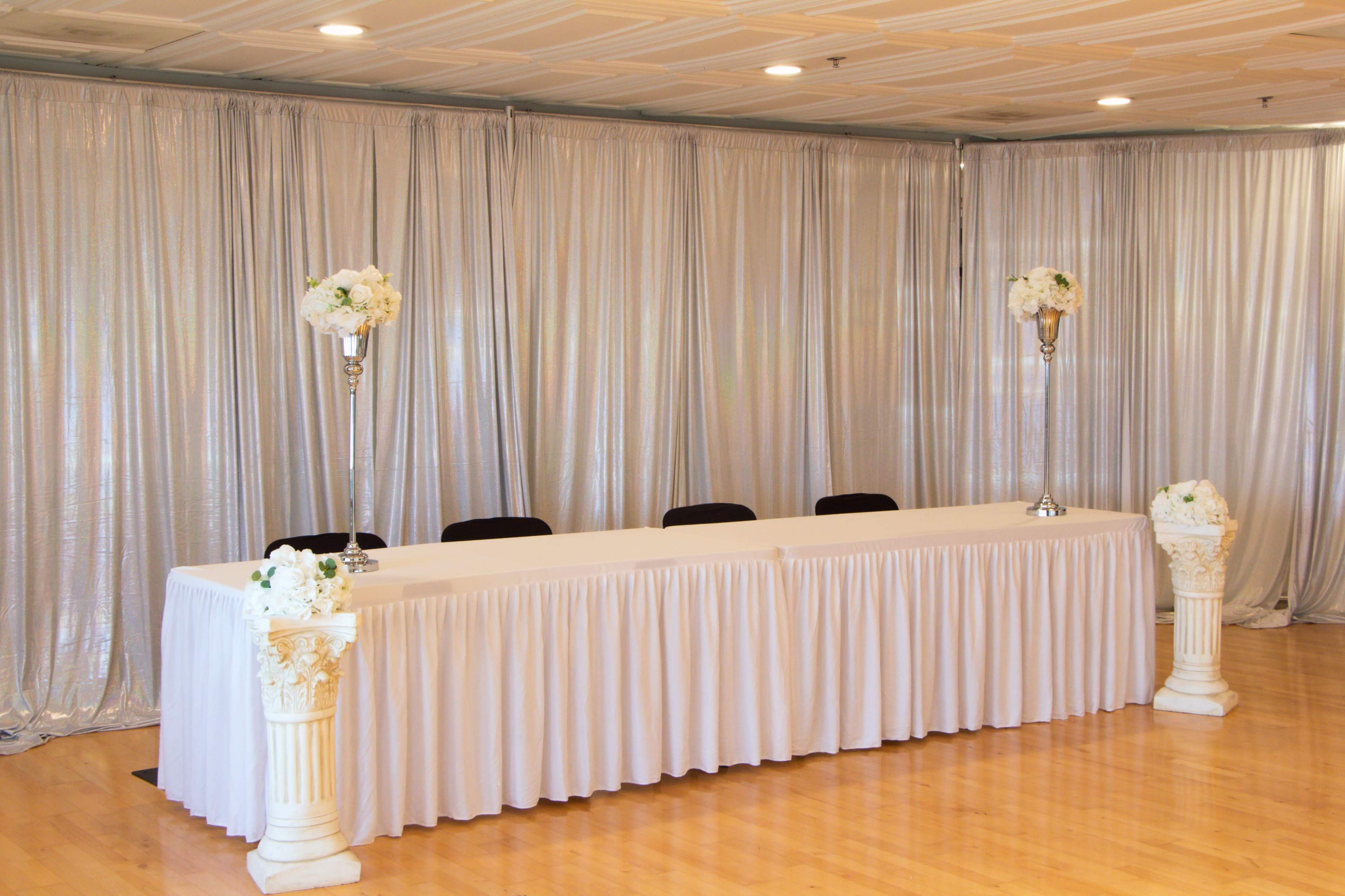 A long white table is set against a backdrop of silver draping, flanked by floral arrangements in vases on tall stands.
