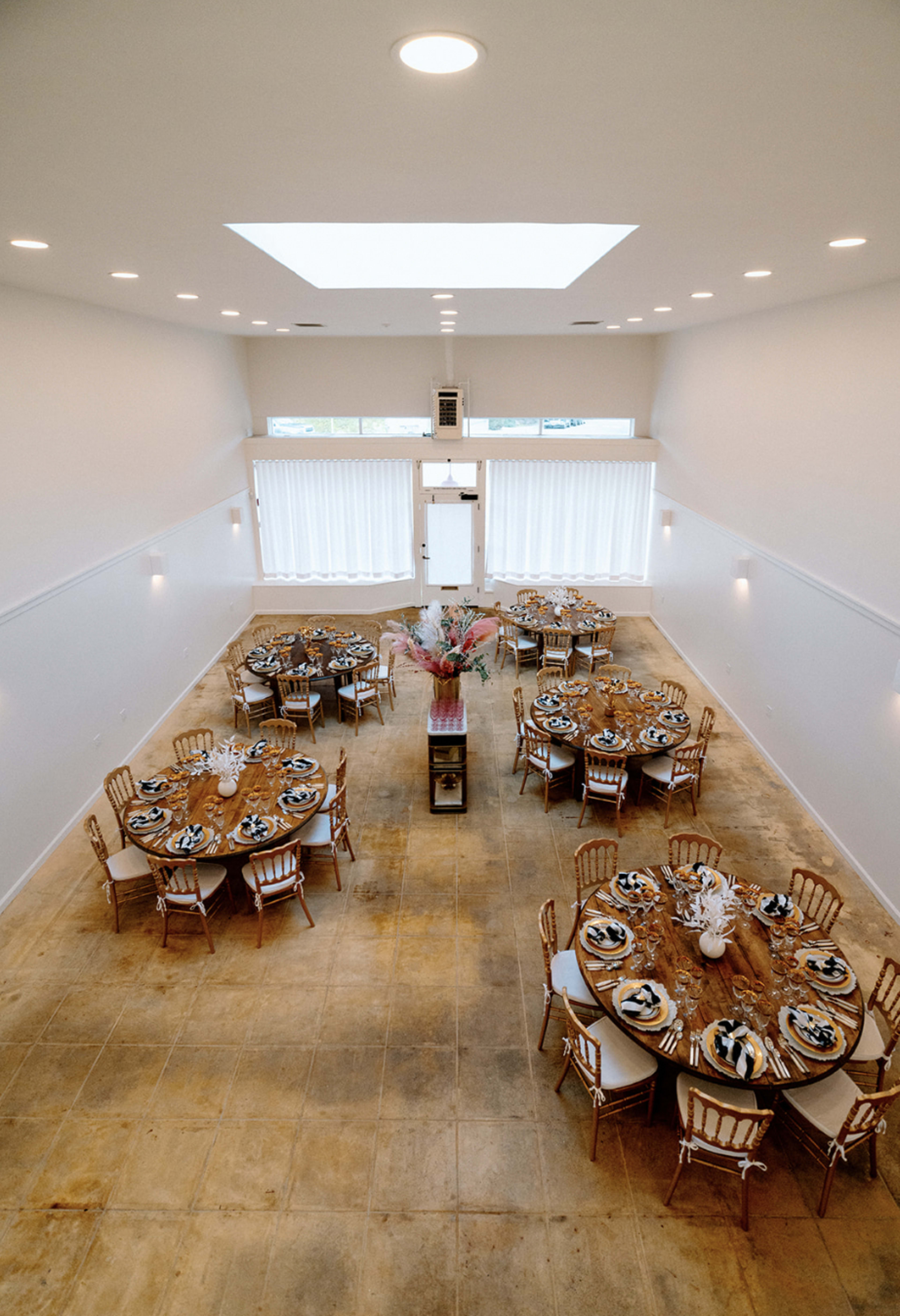 An elegantly arranged event space featuring multiple round tables set for a formal gathering, with large windows allowing natural light to enter.