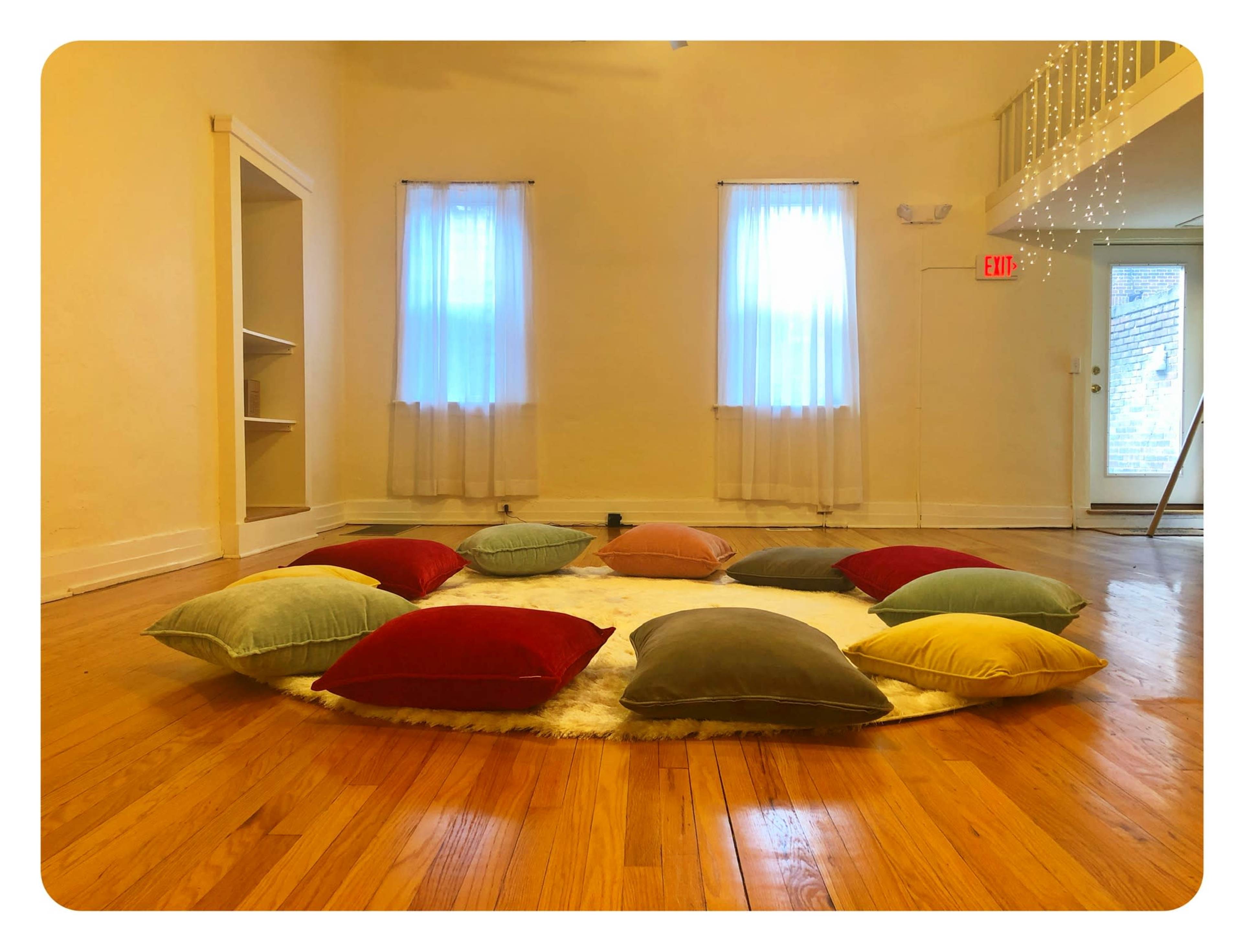 A circular arrangement of colorful pillows on a circular rug in a bright, spacious room with two windows and wooden flooring.
