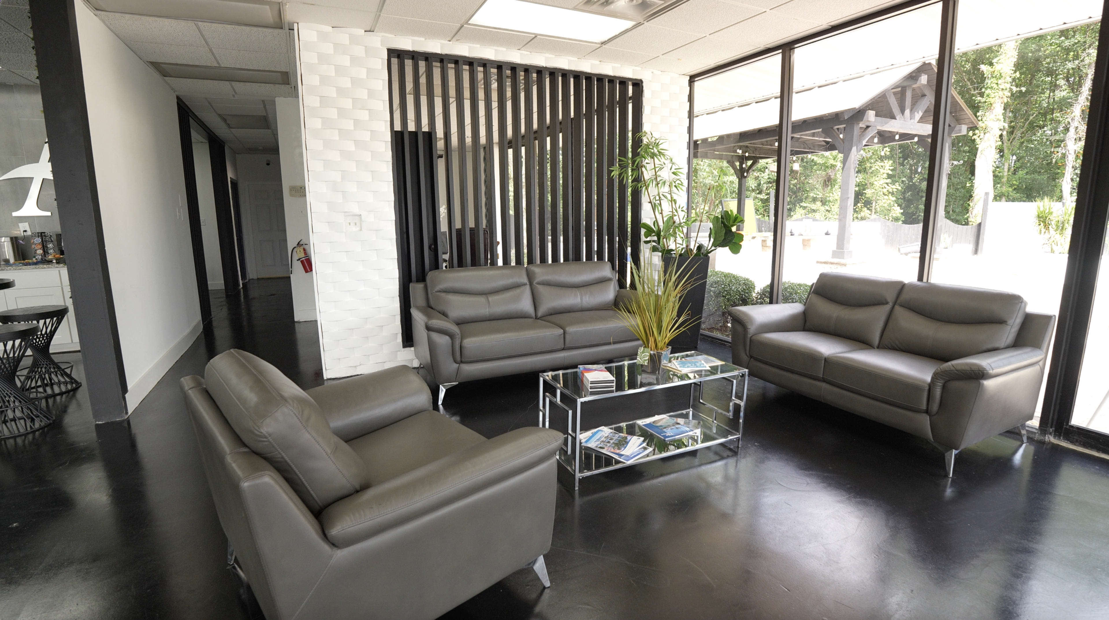 The image shows a modern waiting area with two leather sofas, a glass coffee table, and large windows allowing natural light to fill the space.