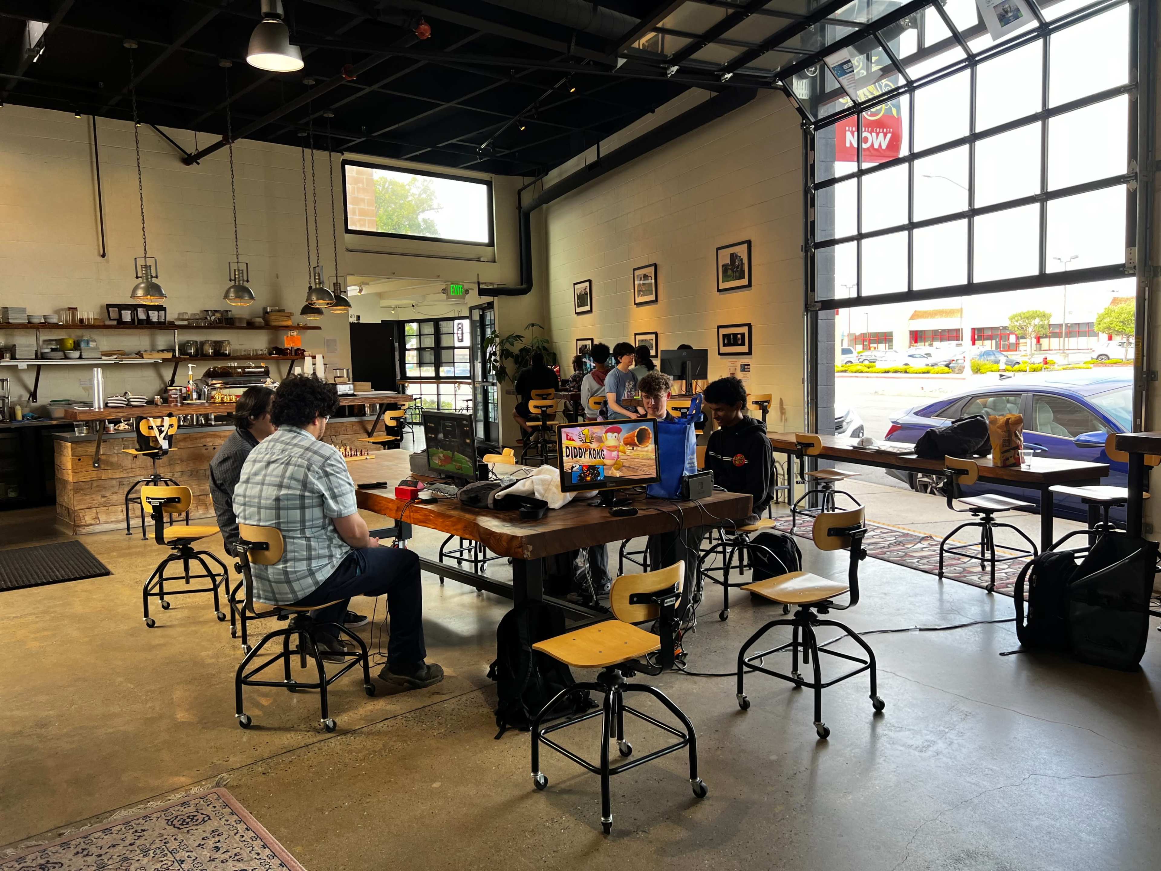 A group of people sits at a large wooden table in a spacious café with large windows and a variety of seating options.