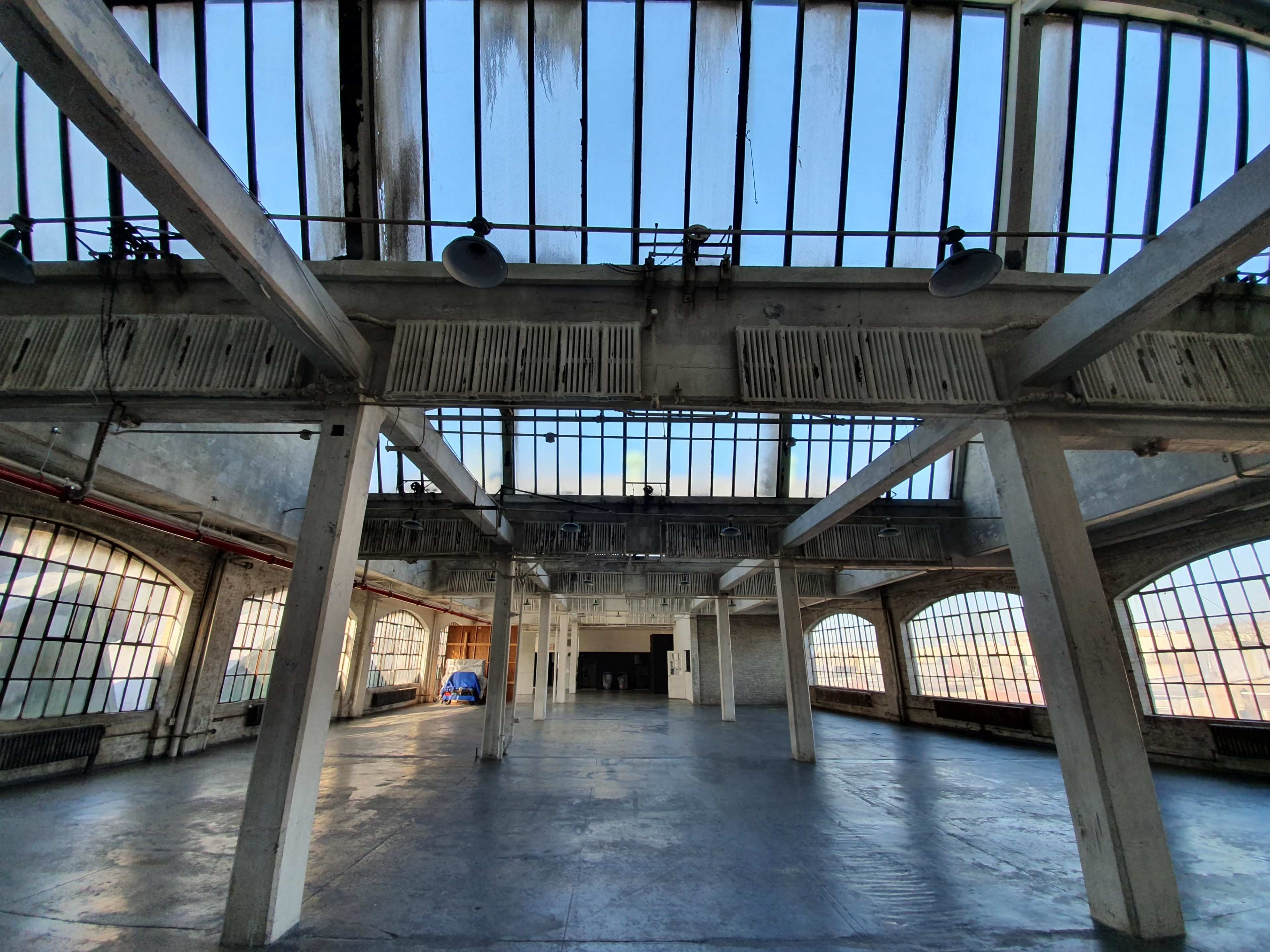 An empty industrial space with large windows and exposed beams, illuminated by natural light.