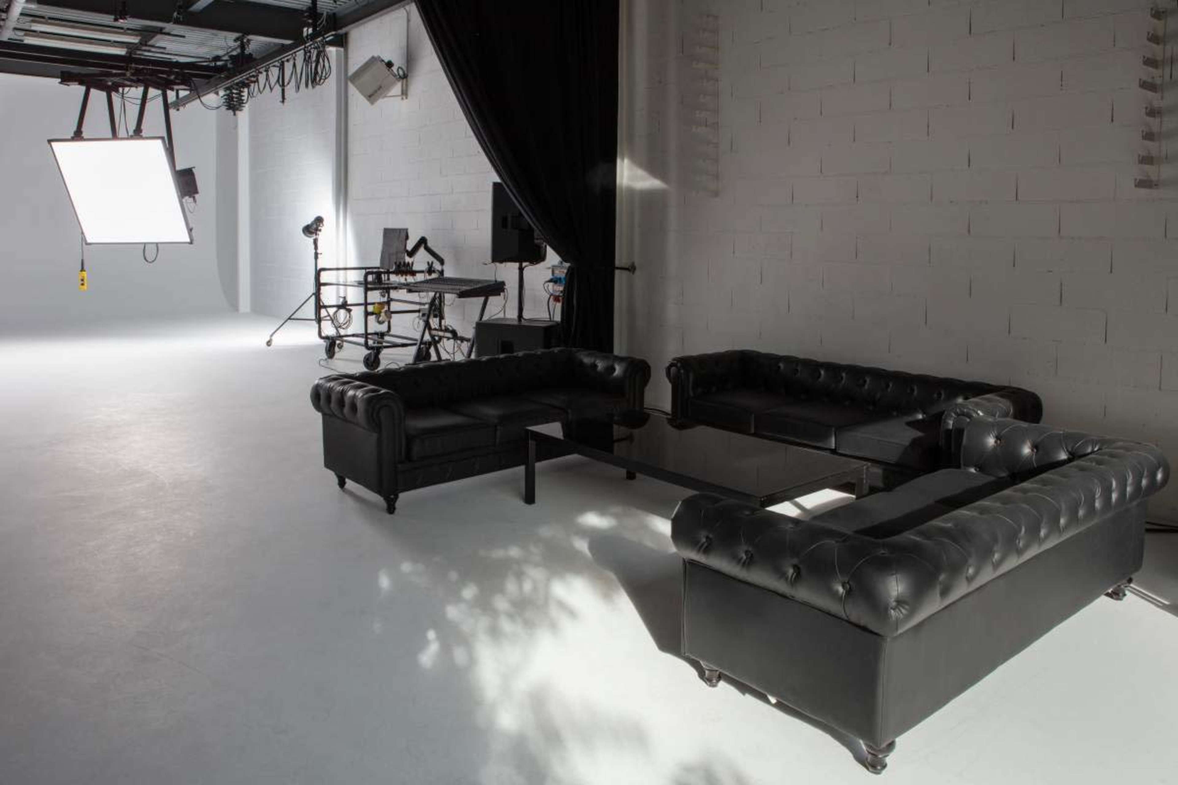 A modern photo studio features black leather couches surrounding a glass table, with professional lighting equipment and backdrops in the background.