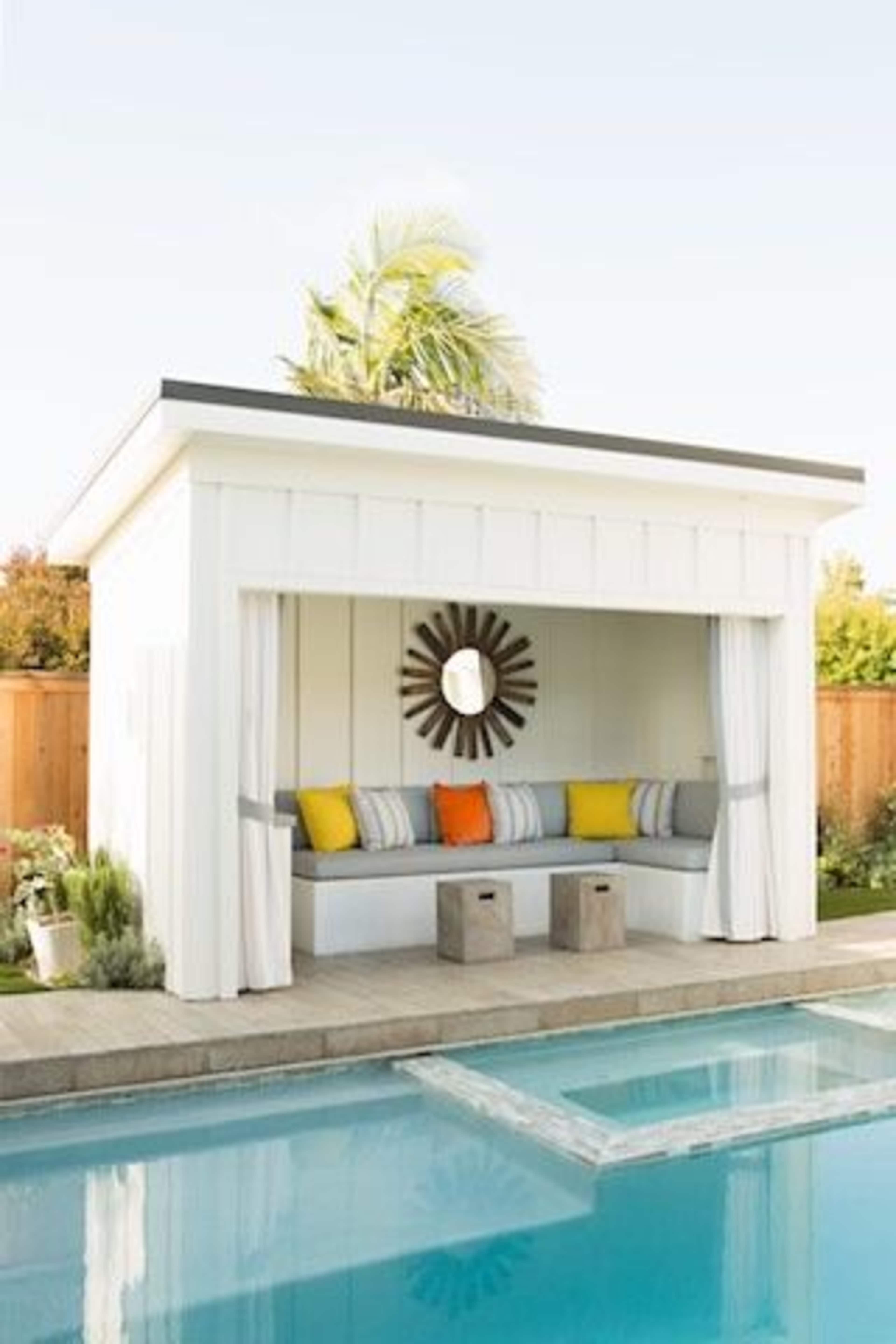A white poolside cabana features a cushioned seating area with colorful pillows, surrounded by greenery and a clear blue pool.