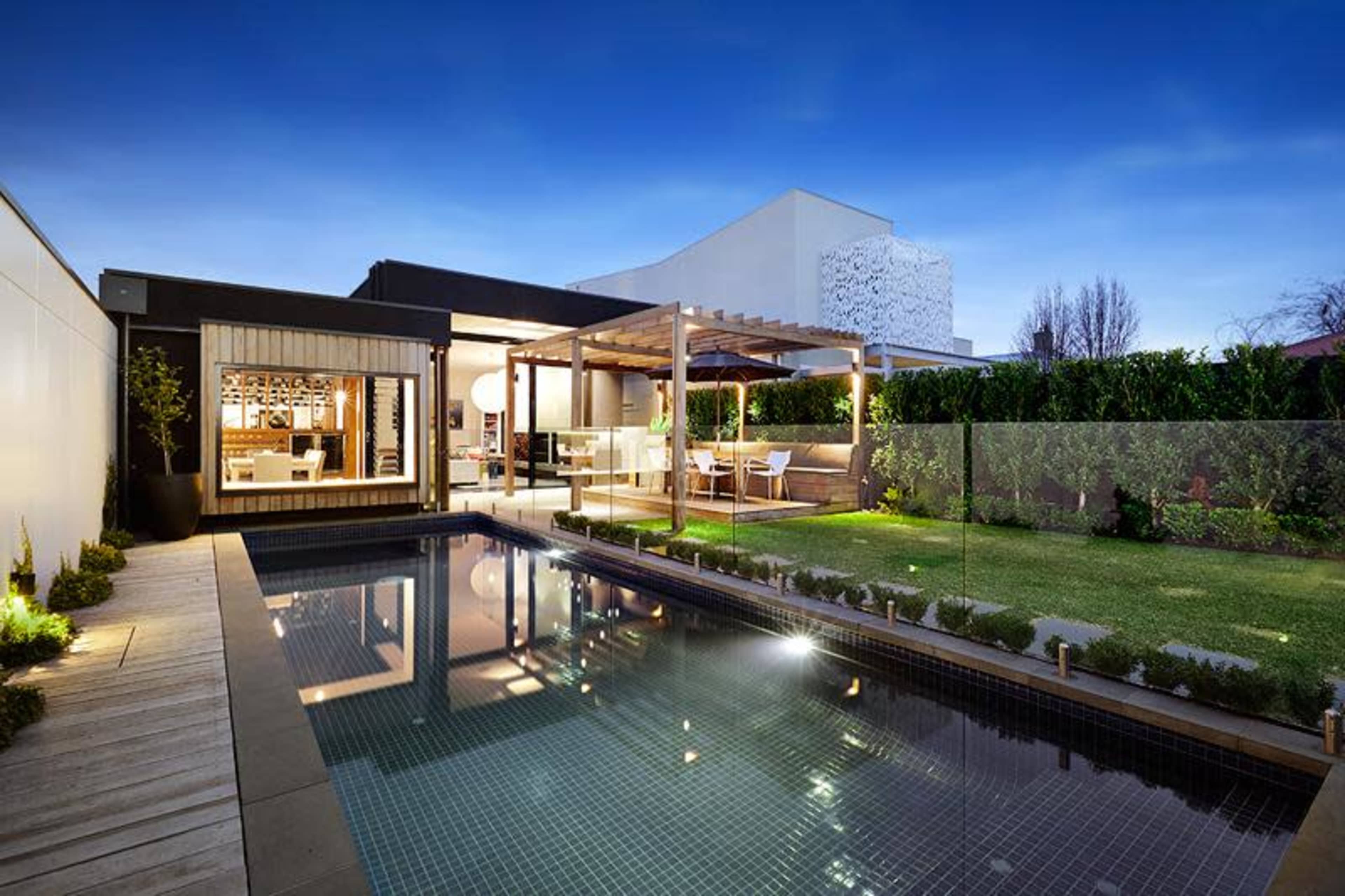 A modern backyard with a swimming pool, wooden deck, and a dining area under a pergola, surrounded by well-maintained greenery at twilight.