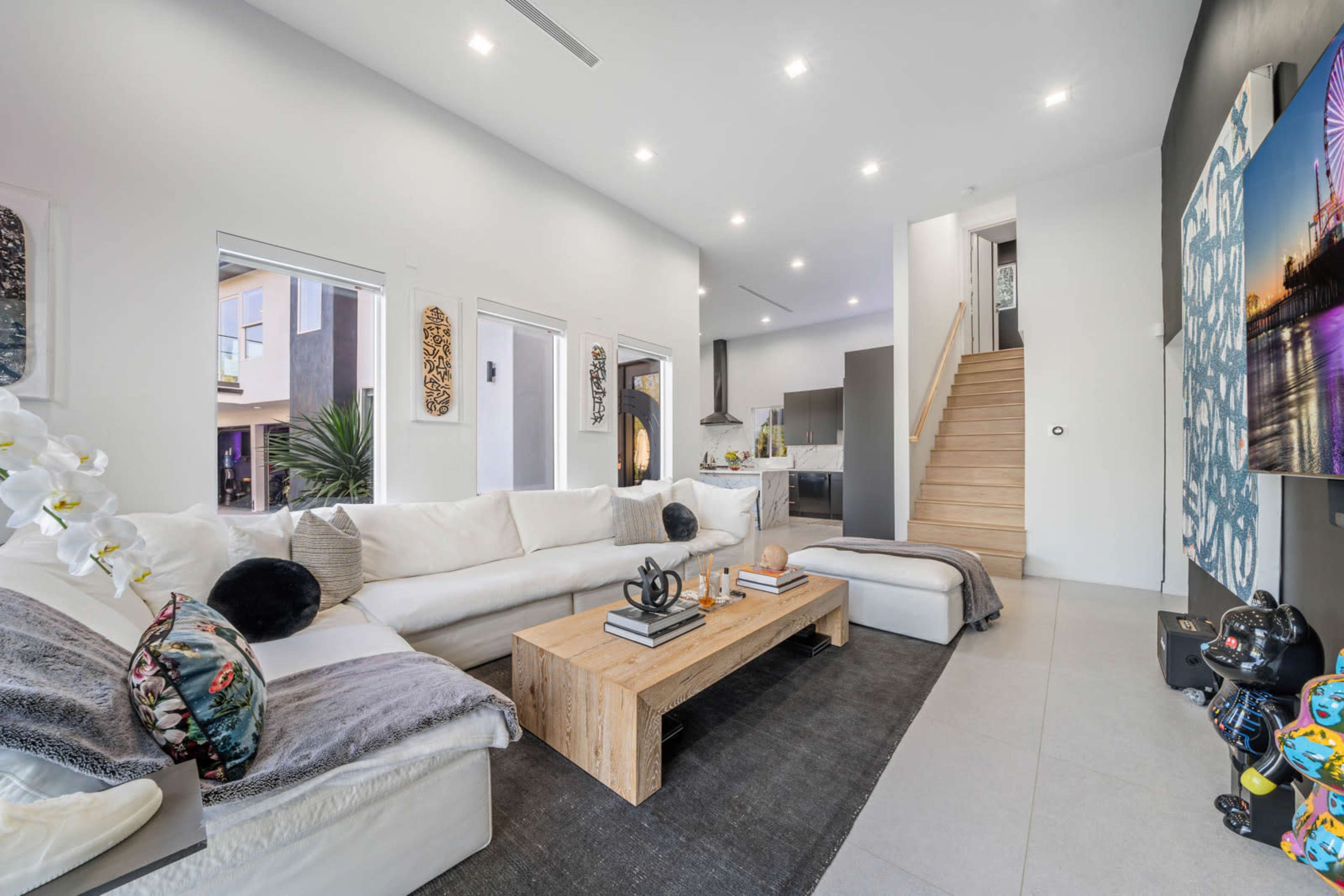 A modern living room features a white sectional sofa, a wooden coffee table, and a staircase leading to an upper level.