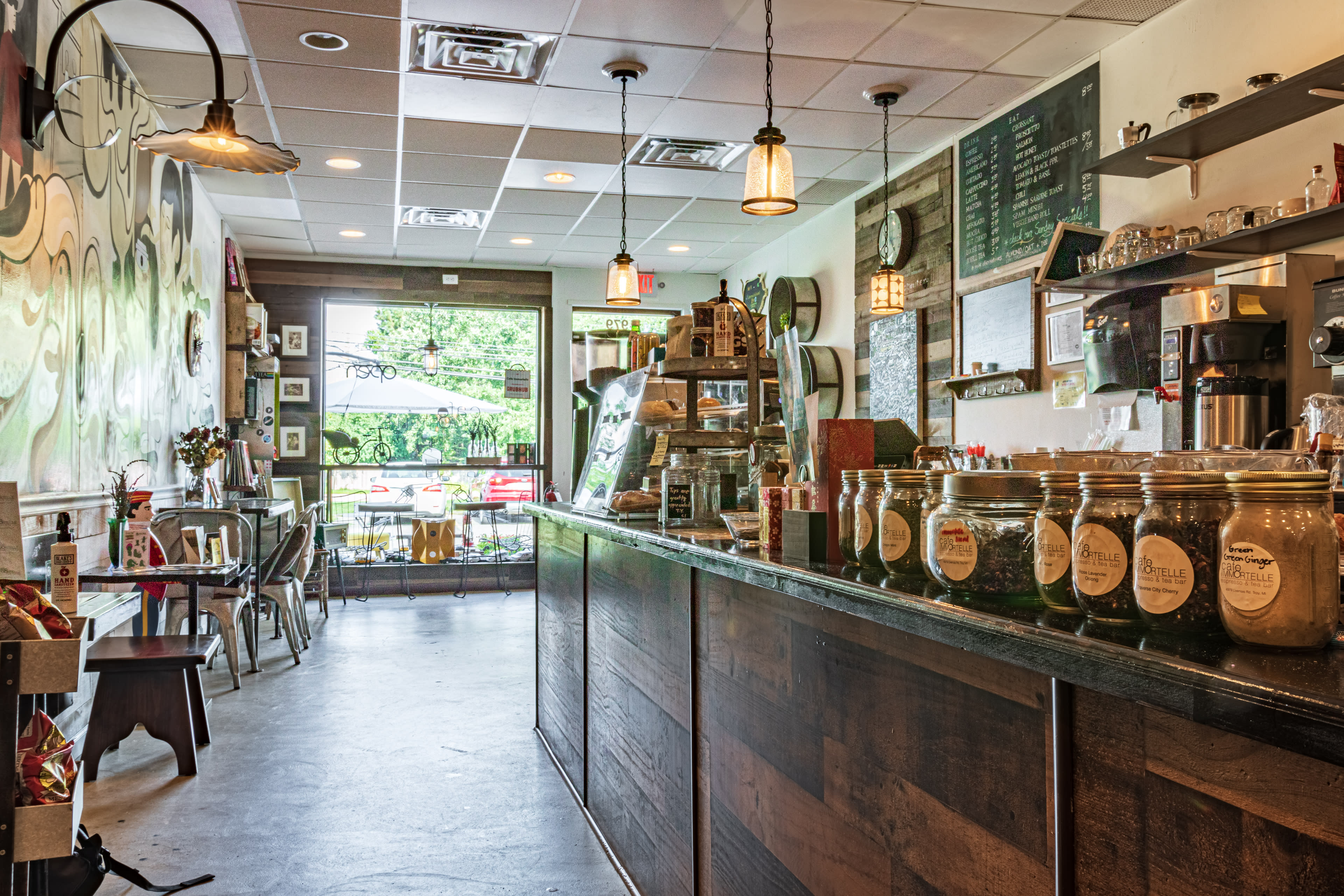 A coffee shop interior features a wooden counter, jars on display, and various seating arrangements, with large windows overlooking an outdoor area.