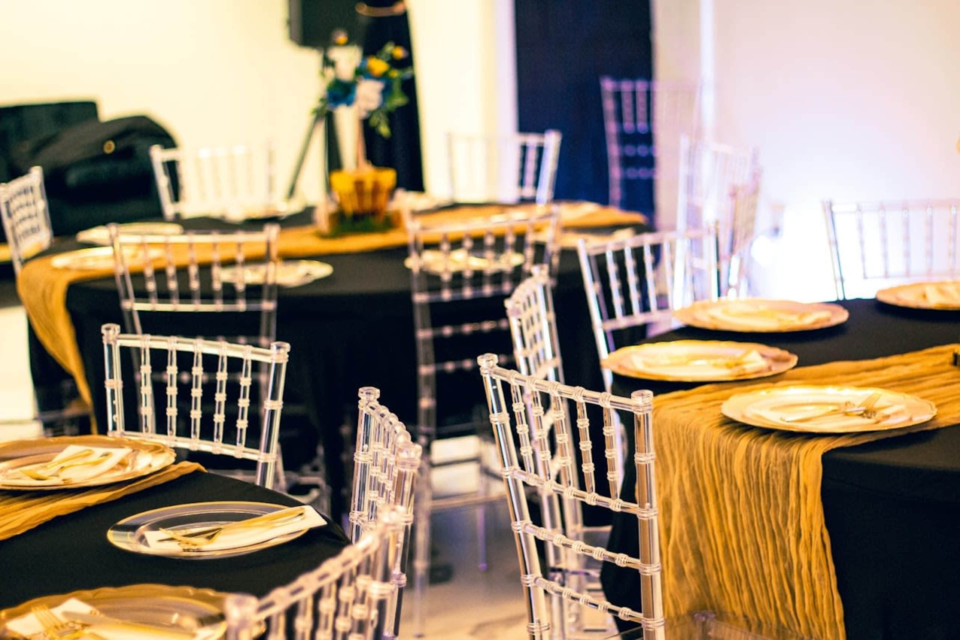 The image shows a banquet setting with clear chairs and black tables adorned with gold table runners and plates.
