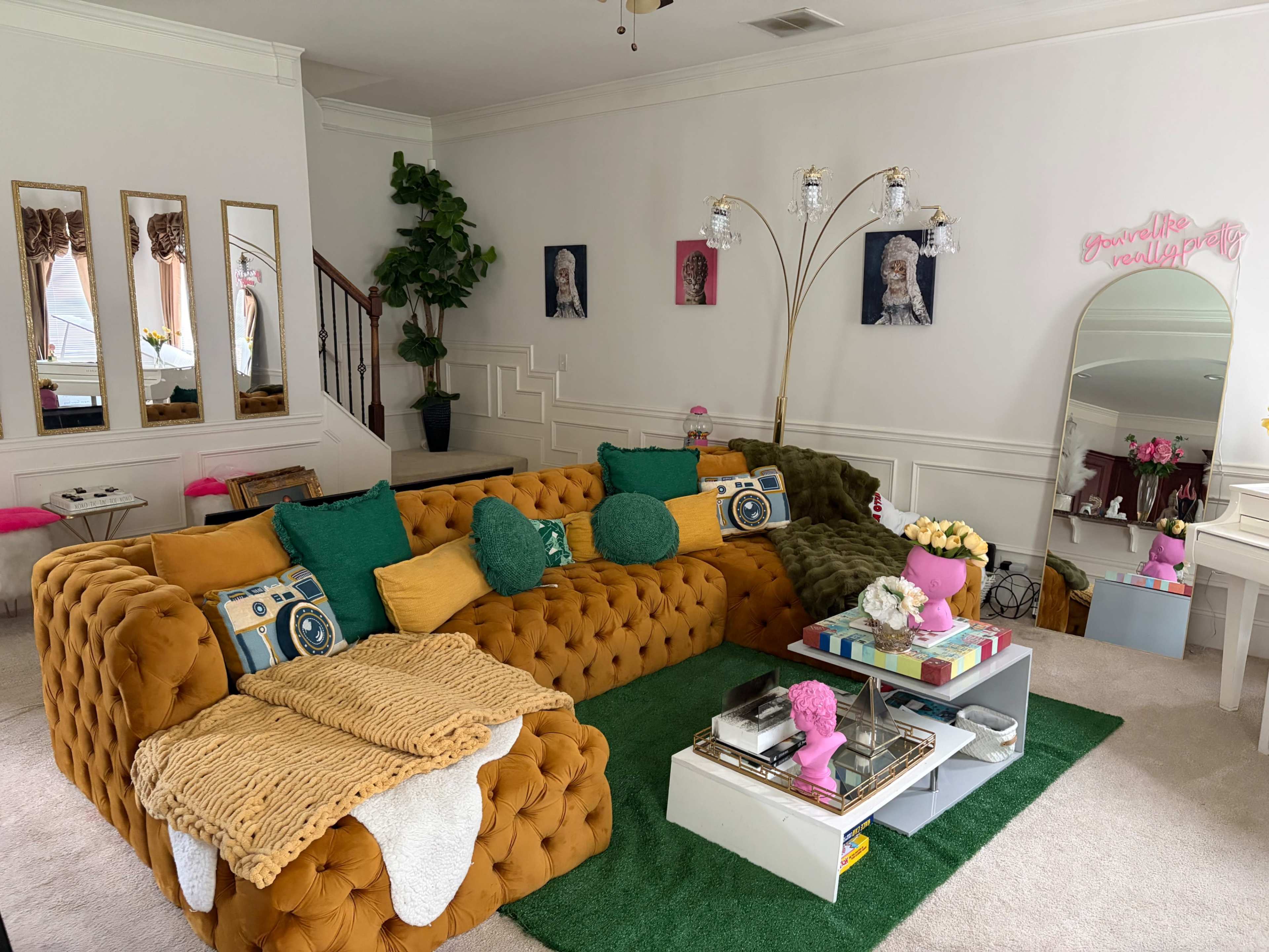 The living room features a large, mustard-colored tufted sofa, a green area rug, and decorative items like a mirror, plants, and art on the walls.