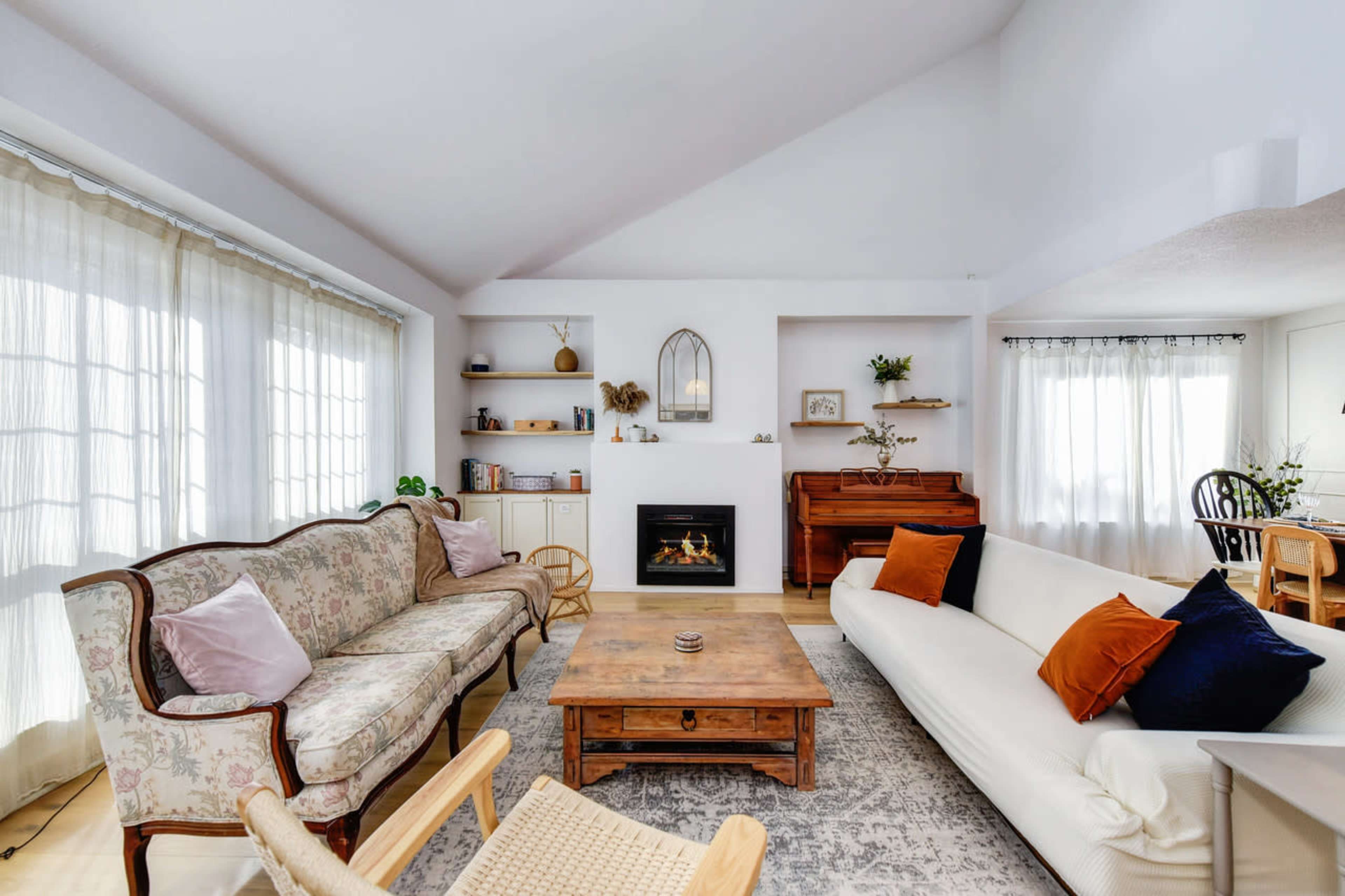 A bright living room features a floral-patterned sofa, a modern white couch, and a wooden coffee table, with a fireplace and a grand piano in the background.