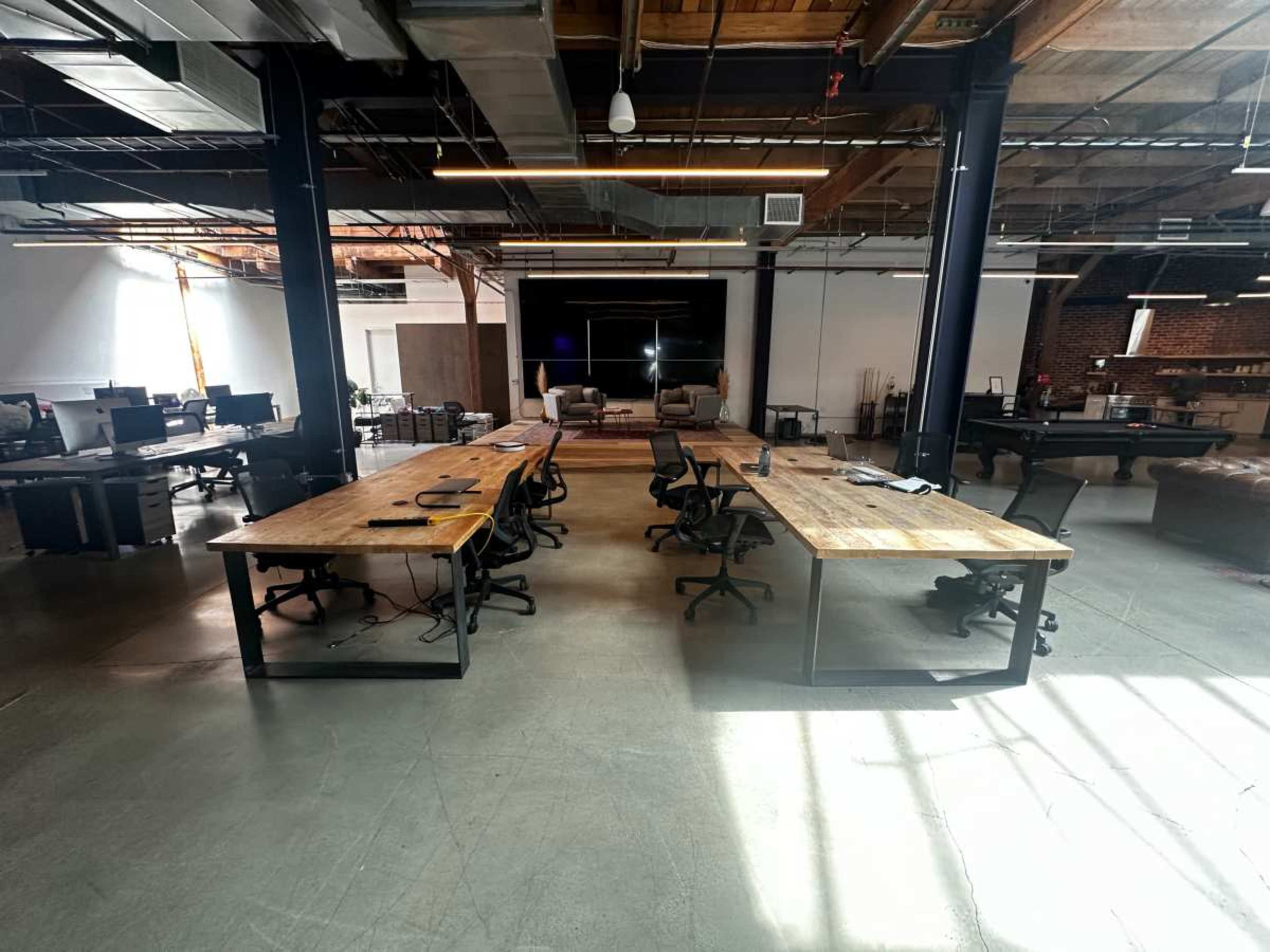 The image shows a modern open office space with large wooden tables, black office chairs, and a few empty workstations under high ceilings and natural light.