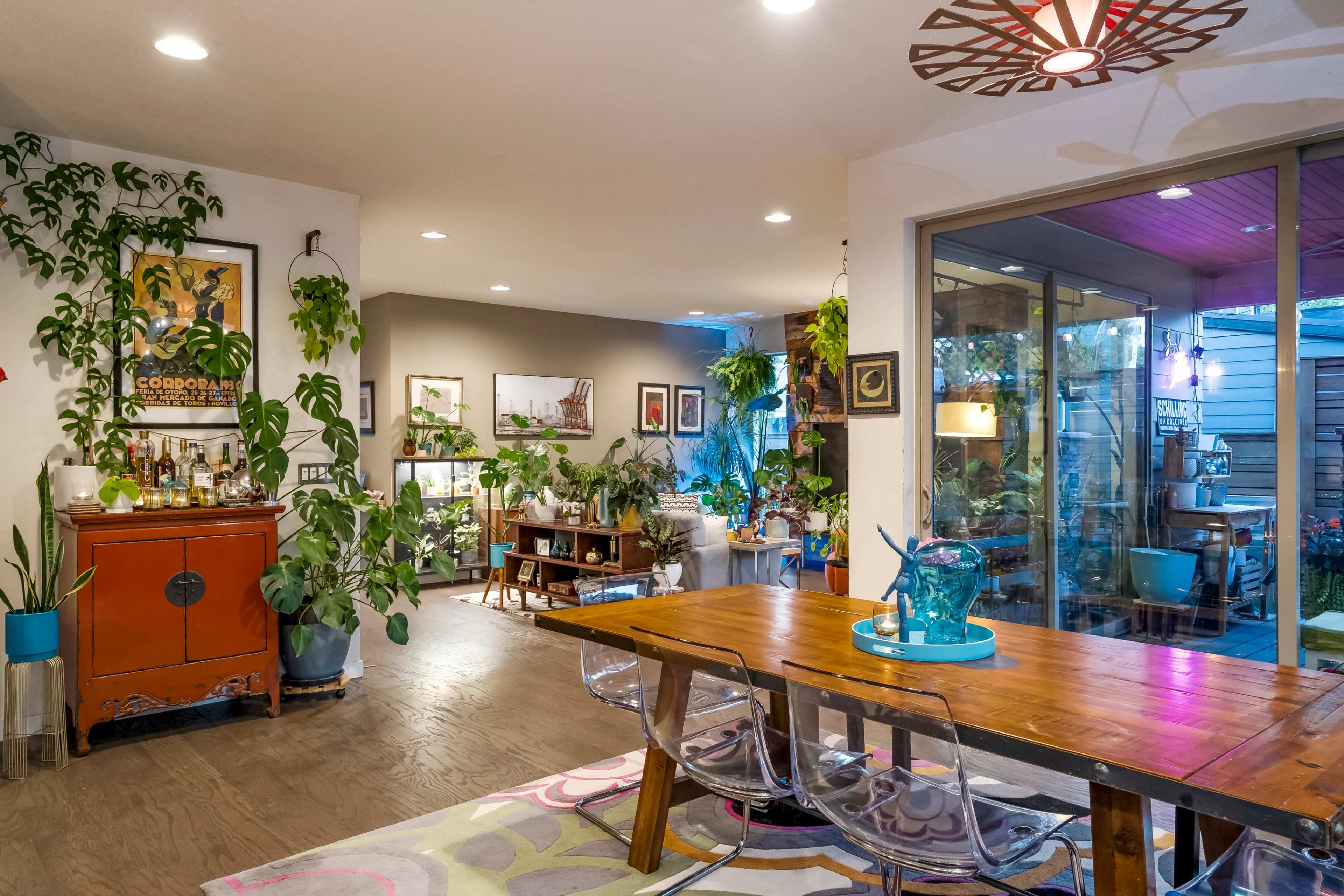 A well-lit living space with a wooden dining table, transparent chairs, and various plants lining the walls, alongside framed artwork and a sliding glass door leading to an outdoor area.