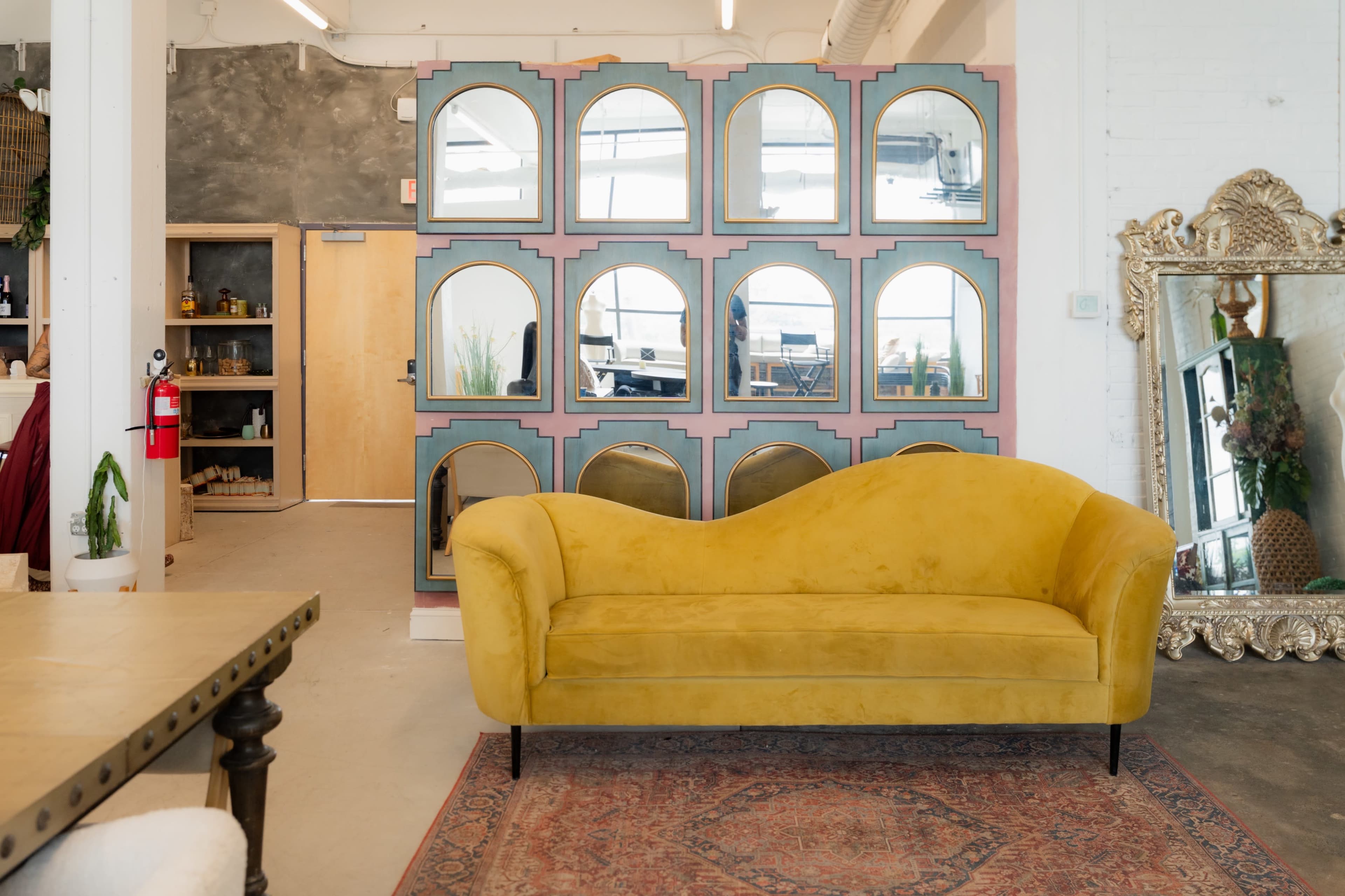 The image features a yellow sofa in front of a decorative mirror wall, with a wooden table and a door leading to another room in the background.