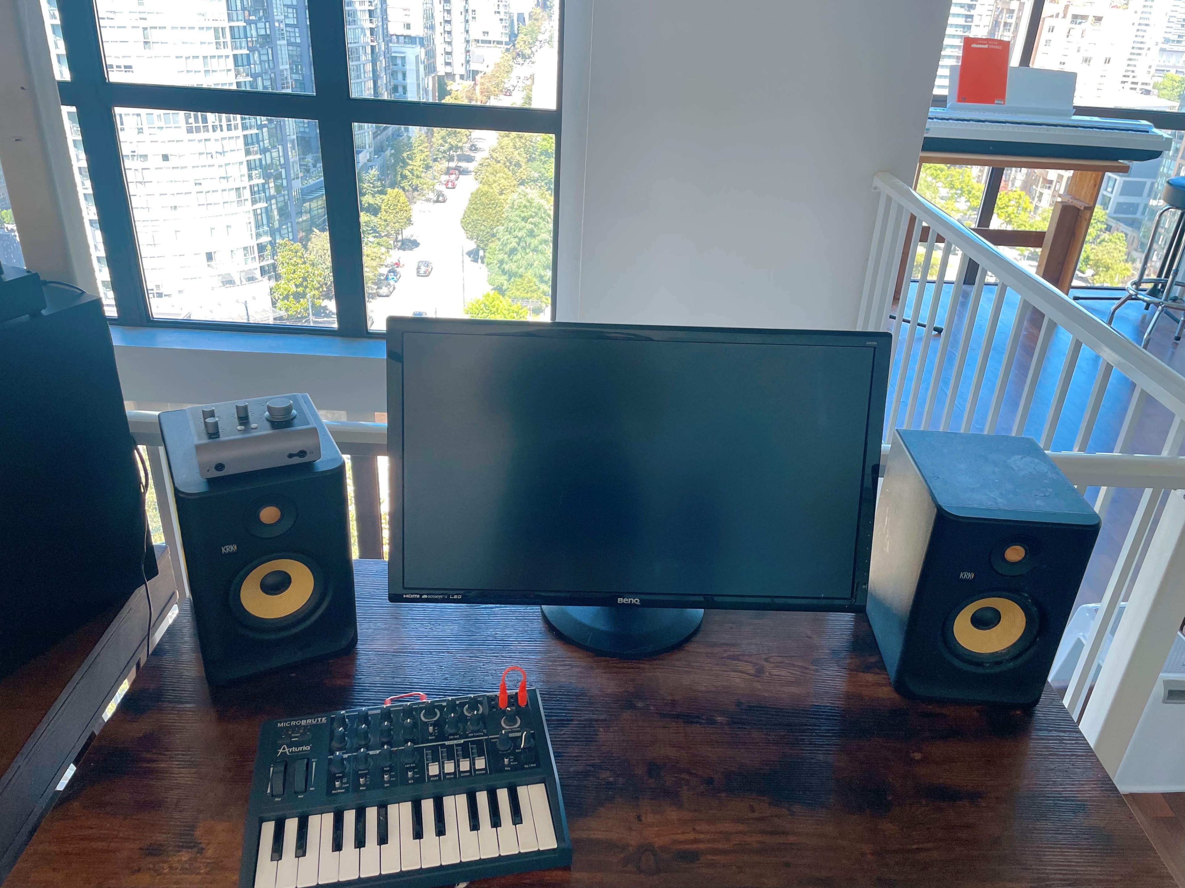 A computer monitor sits on a wooden desk flanked by two speakers and a MIDI keyboard, with a view of a city street visible through large windows.