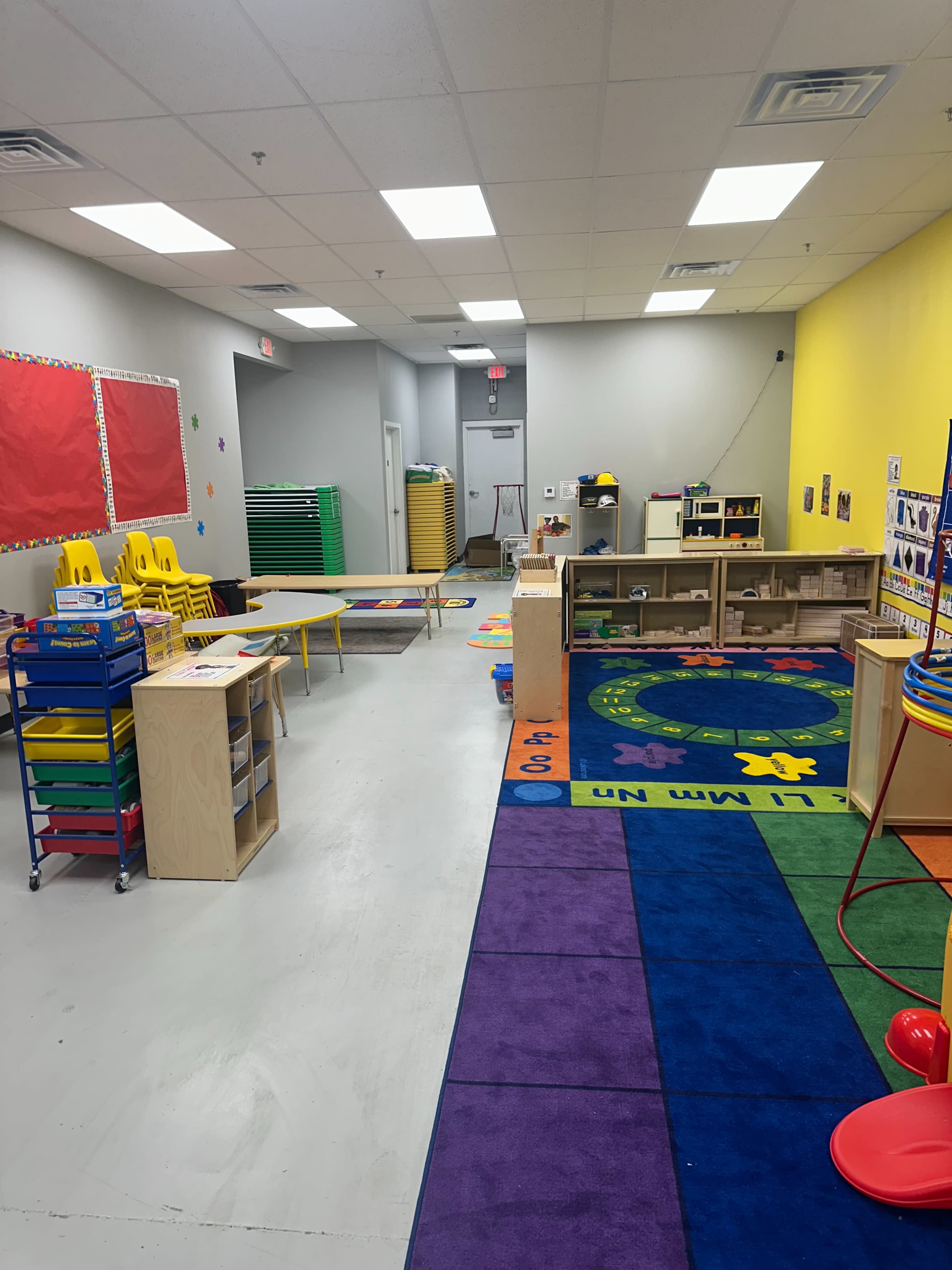 The image shows a colorful kindergarten classroom with yellow chairs, educational toys, and a bright rug, arranged along gray walls.