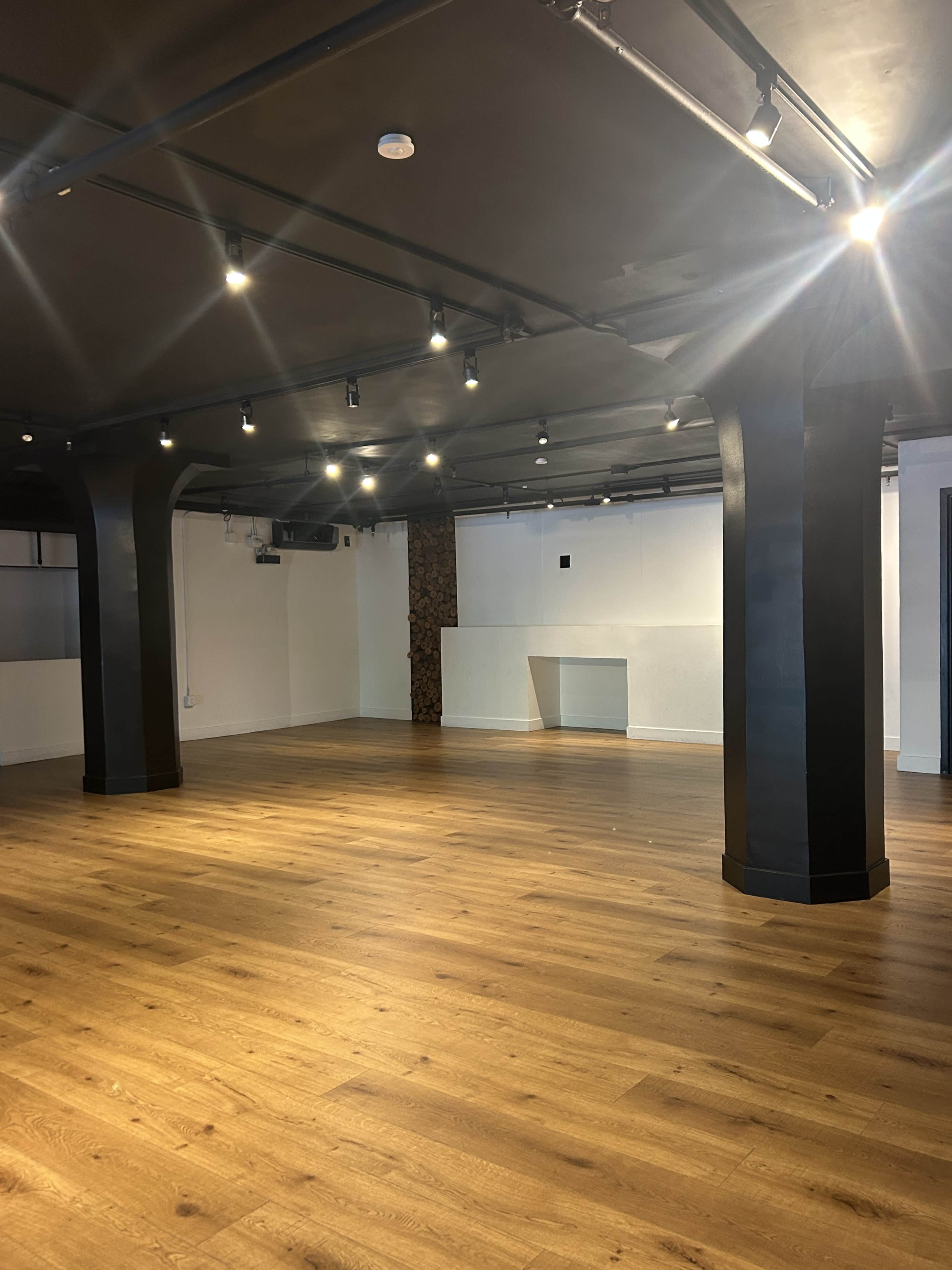 The image shows a spacious room with wooden flooring, black columns, and overhead lights illuminating the area.