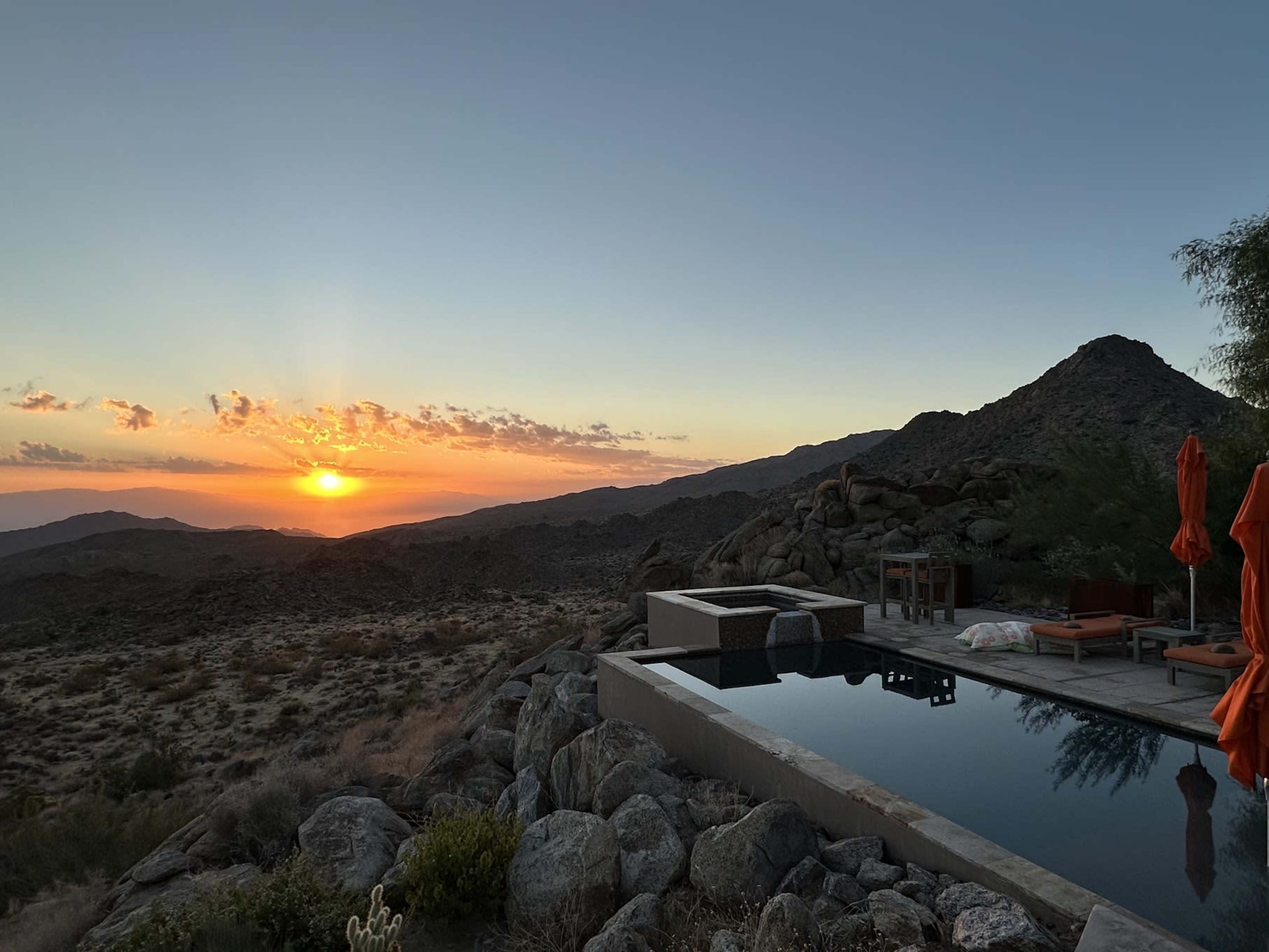 A swimming pool with lounge chairs is situated in a rocky landscape as the sun sets behind the mountains.